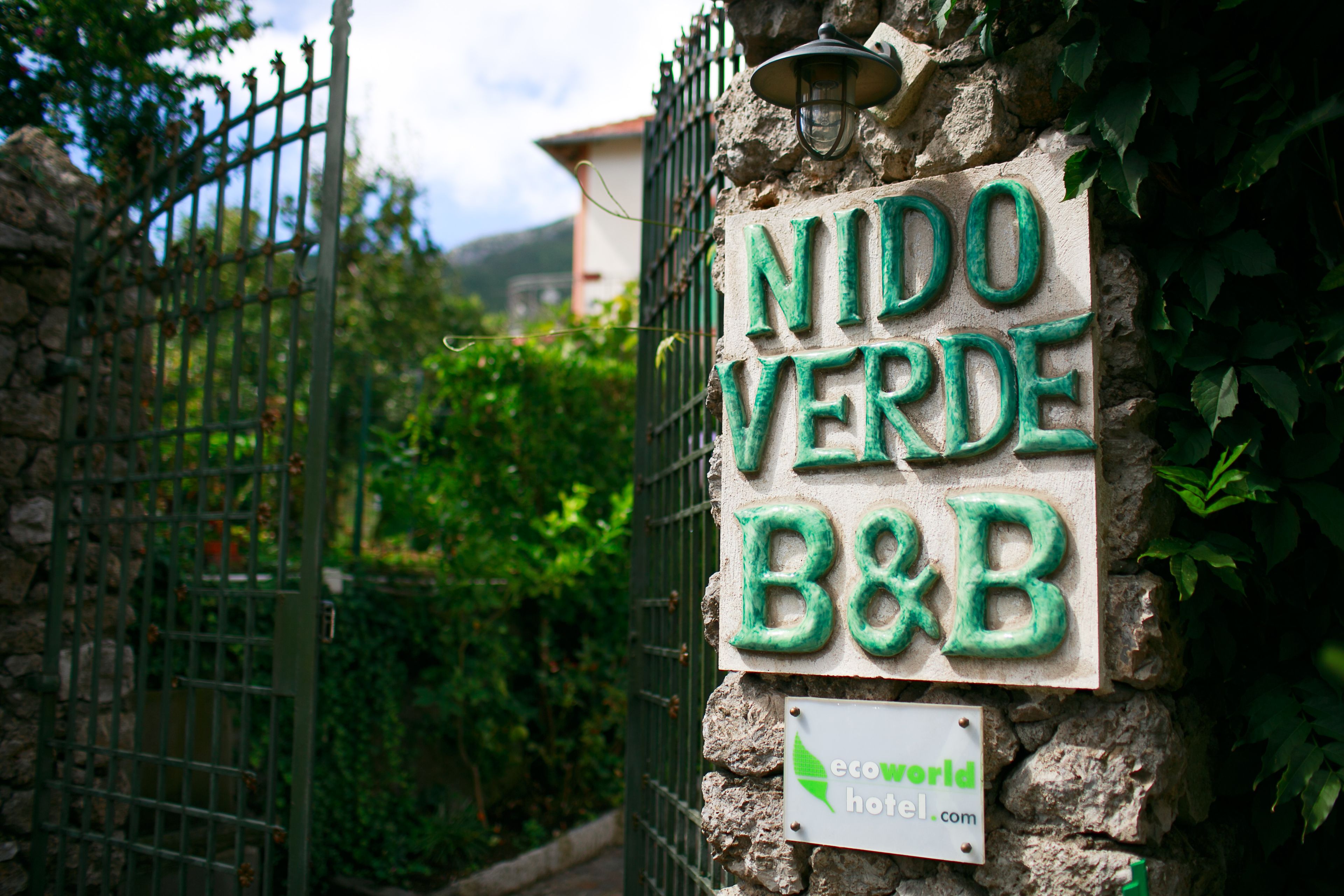 Un mur de pierre sur lequel est apposée une enseigne indiquant « NIDO VERDE B&B », et en dessous, une plus petite enseigne portant le nom de domaine « ecoworld hotel.com ».