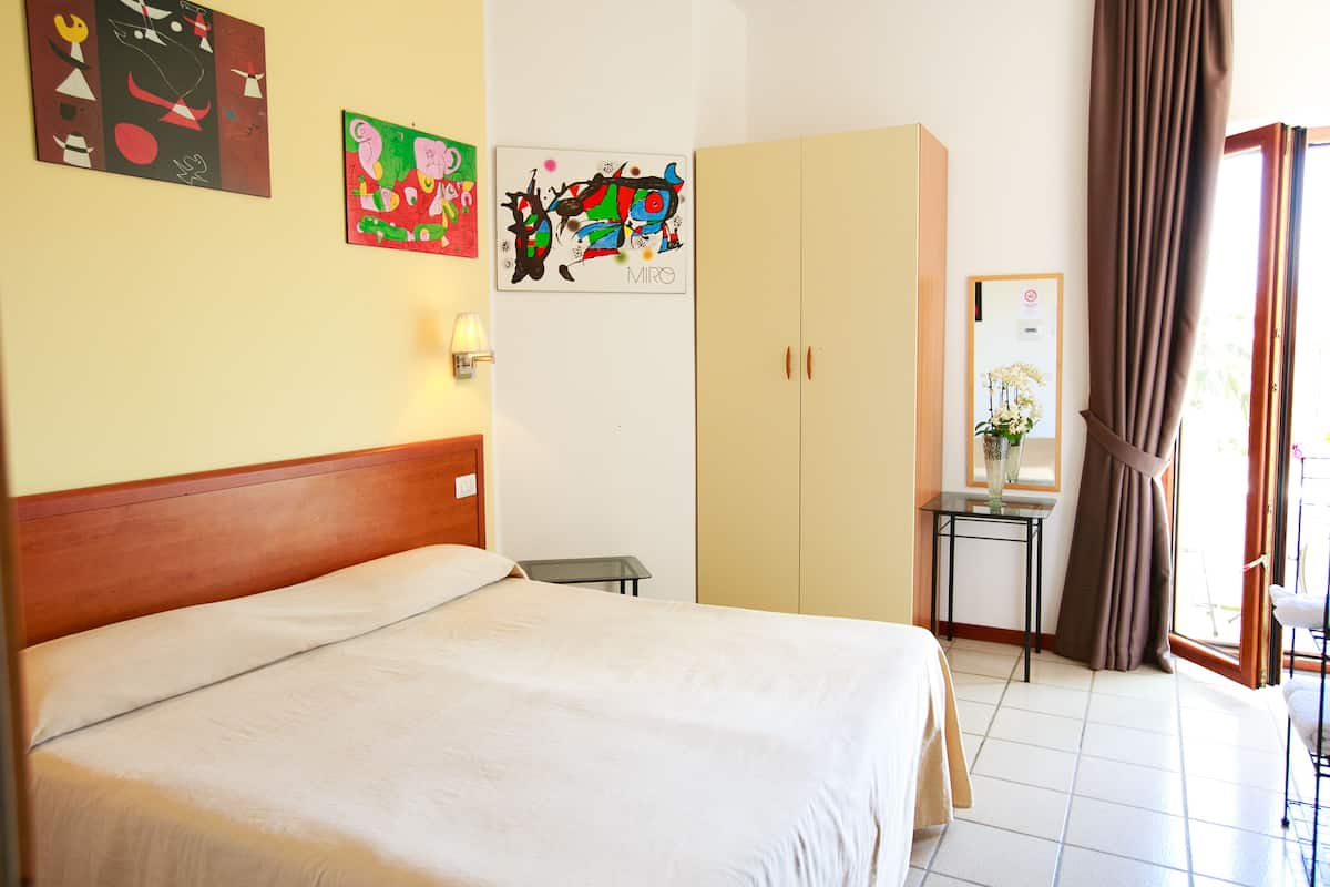 A hotel room with a bed, a wardrobe, a small table with a vase, and artwork on the walls.
