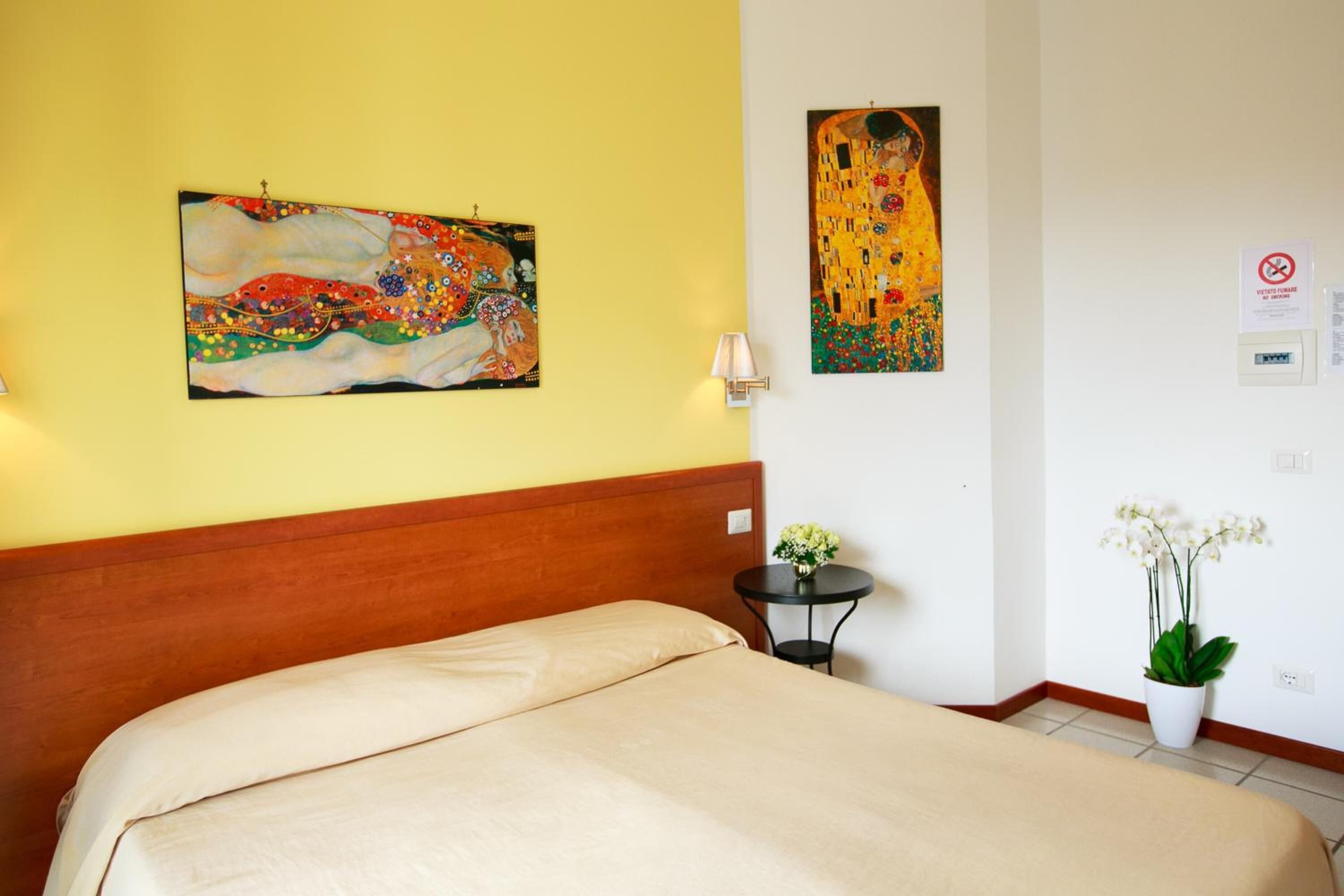 Une chambre d’hôtel avec un lit, un mur orné de deux tableaux, une petite table avec une lampe et une plante en pot.