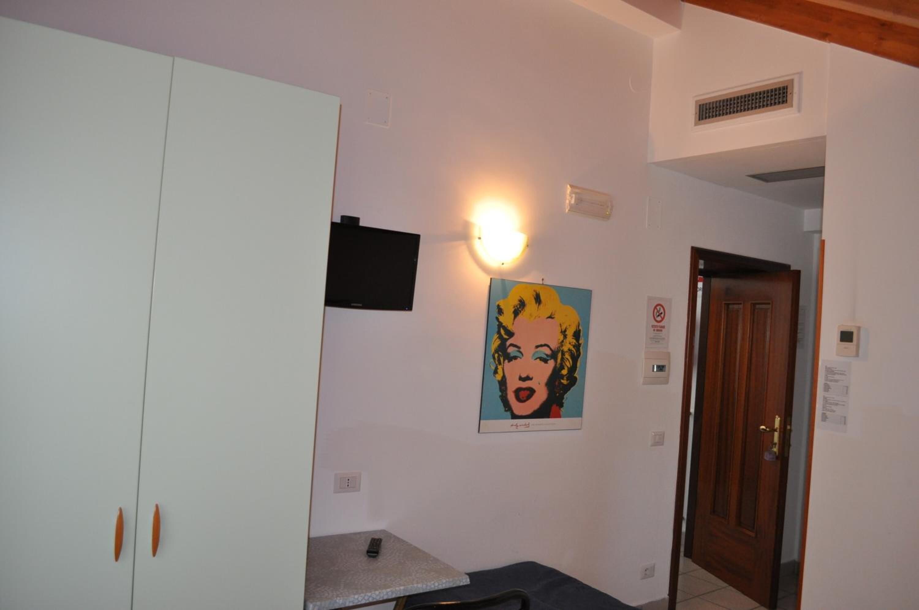 A room with a white wardrobe, a small table, a wall-mounted light, and a door.