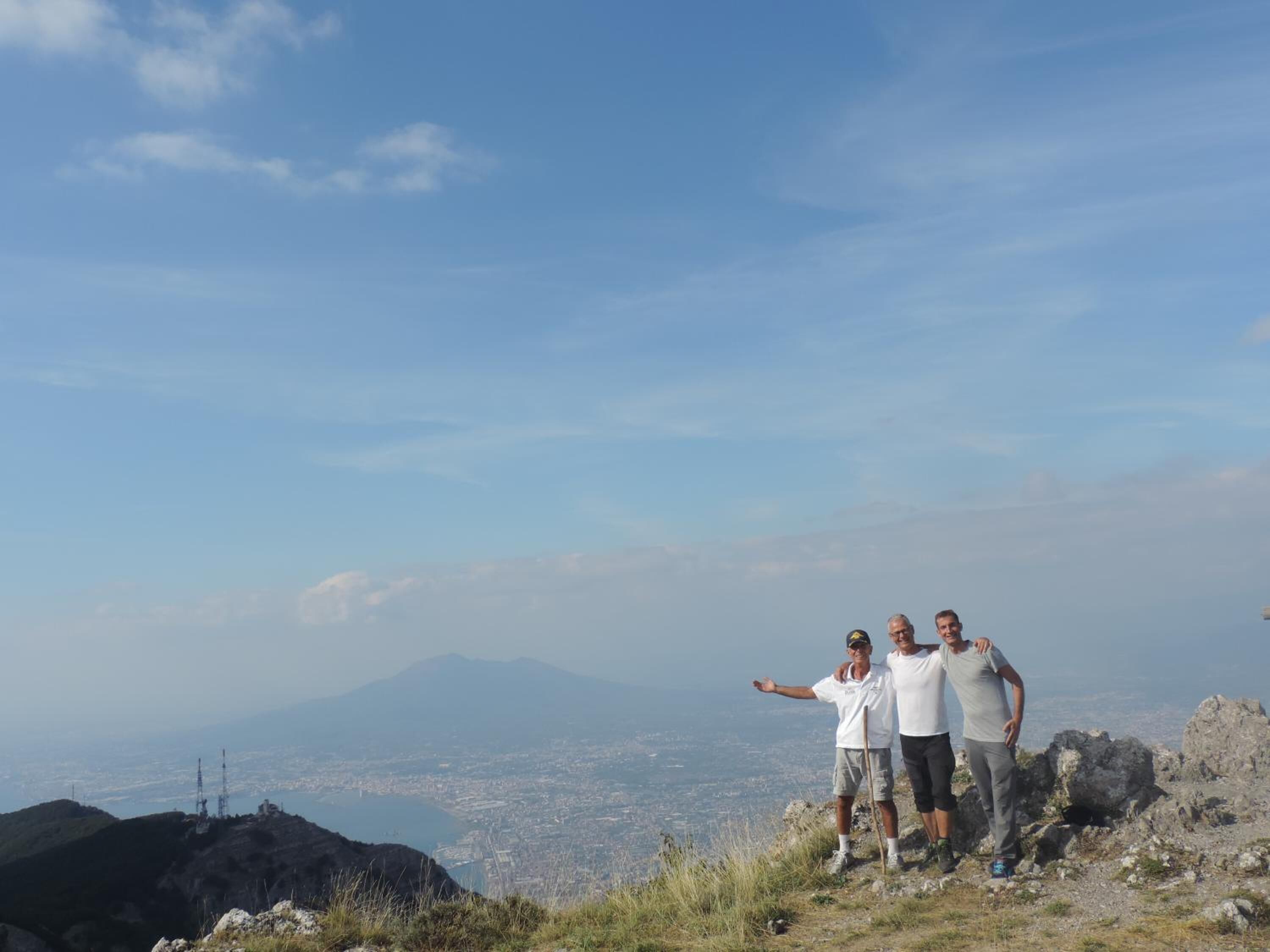 Three people standing on a rocky hilltop with a cityscape and mountains in the background.