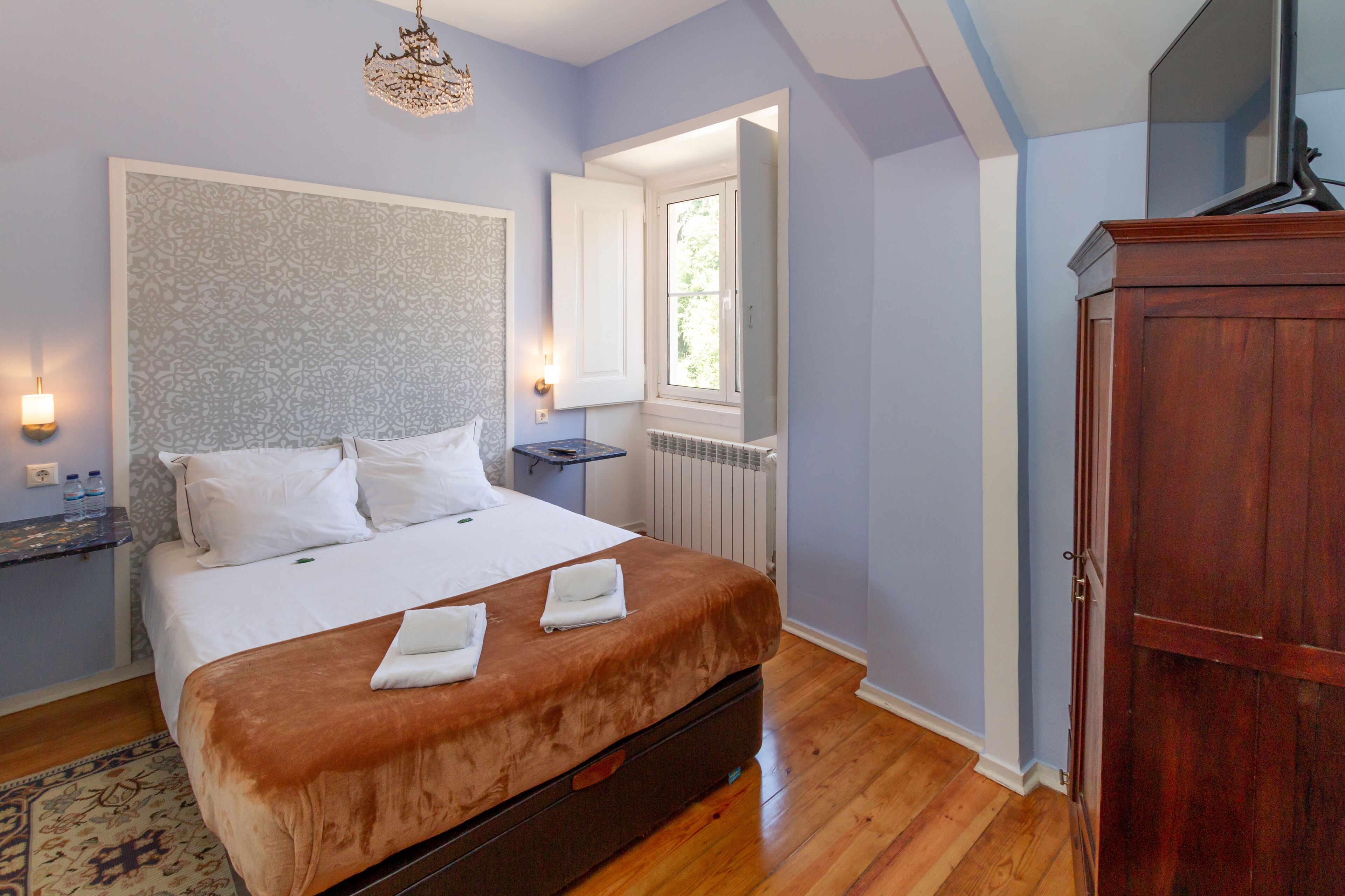 A bedroom with a large bed, a wooden wardrobe, a nightstand, and a window with a view of greenery.
