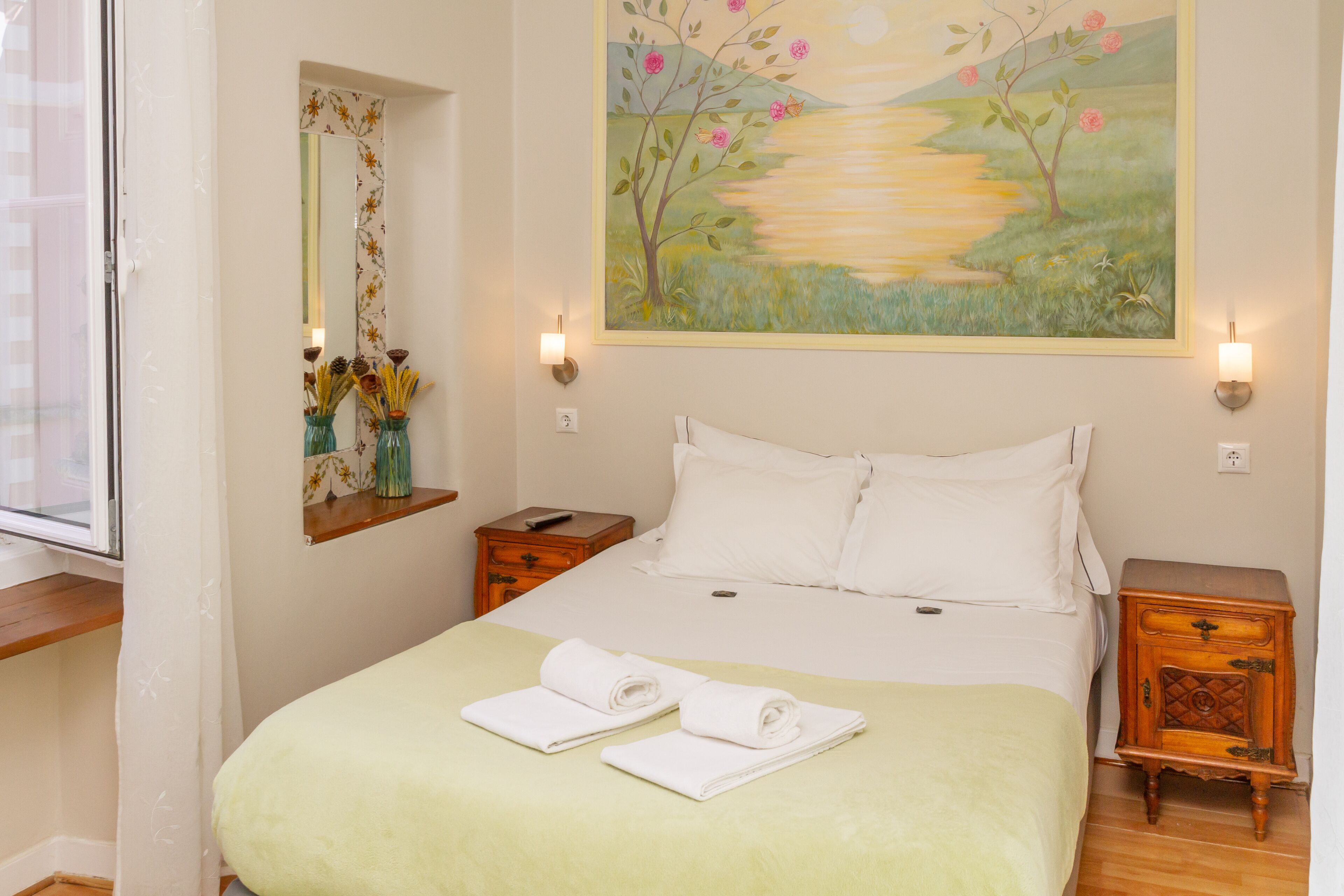 A neatly made bed with white linens and two rolled towels, a painting of a landscape on the wall, and a window with a view of the outdoors.