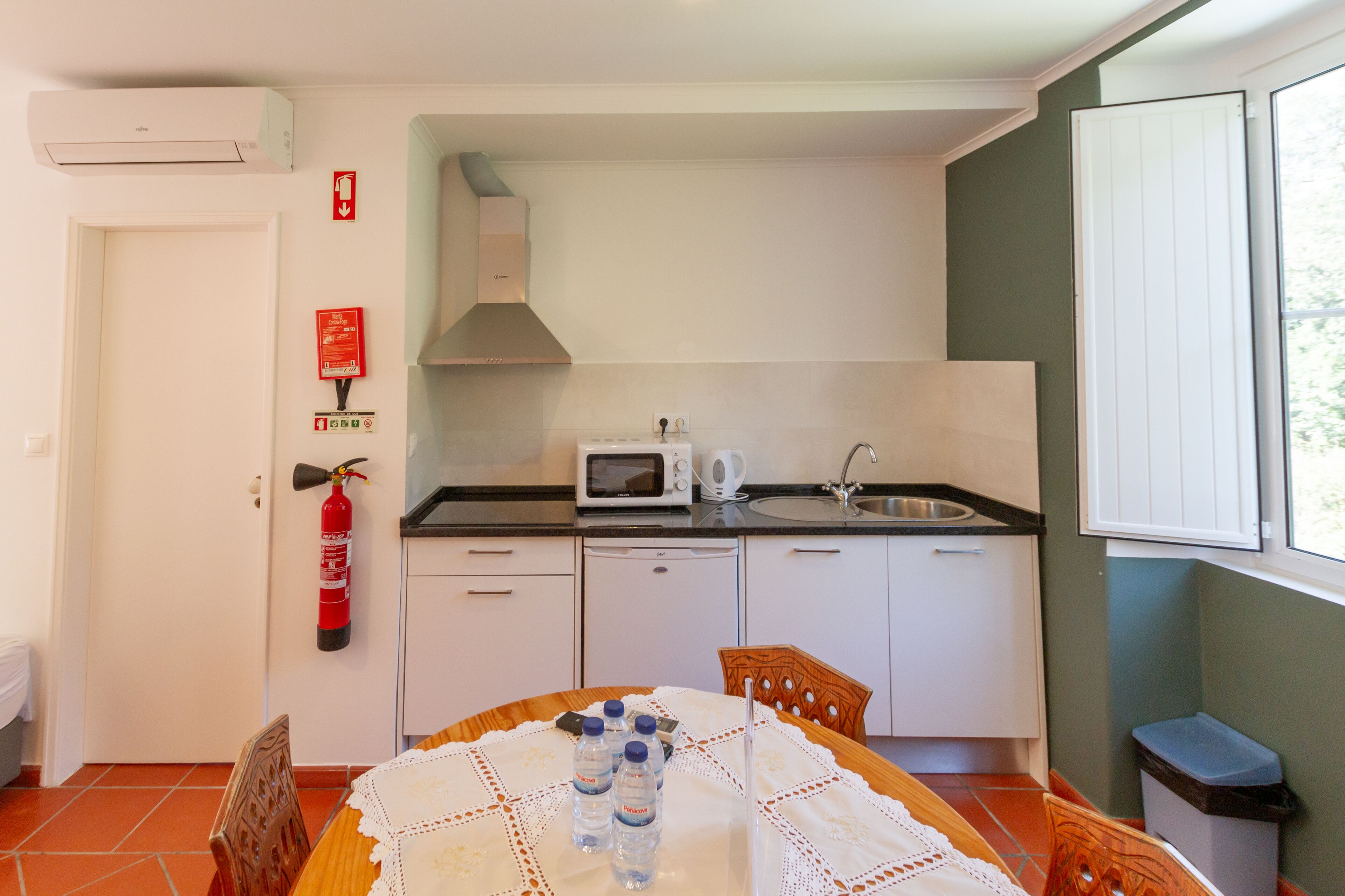 A compact kitchen with a microwave, sink, and white cabinets. There is a fire extinguisher on the wall and a window with a view of greenery.
