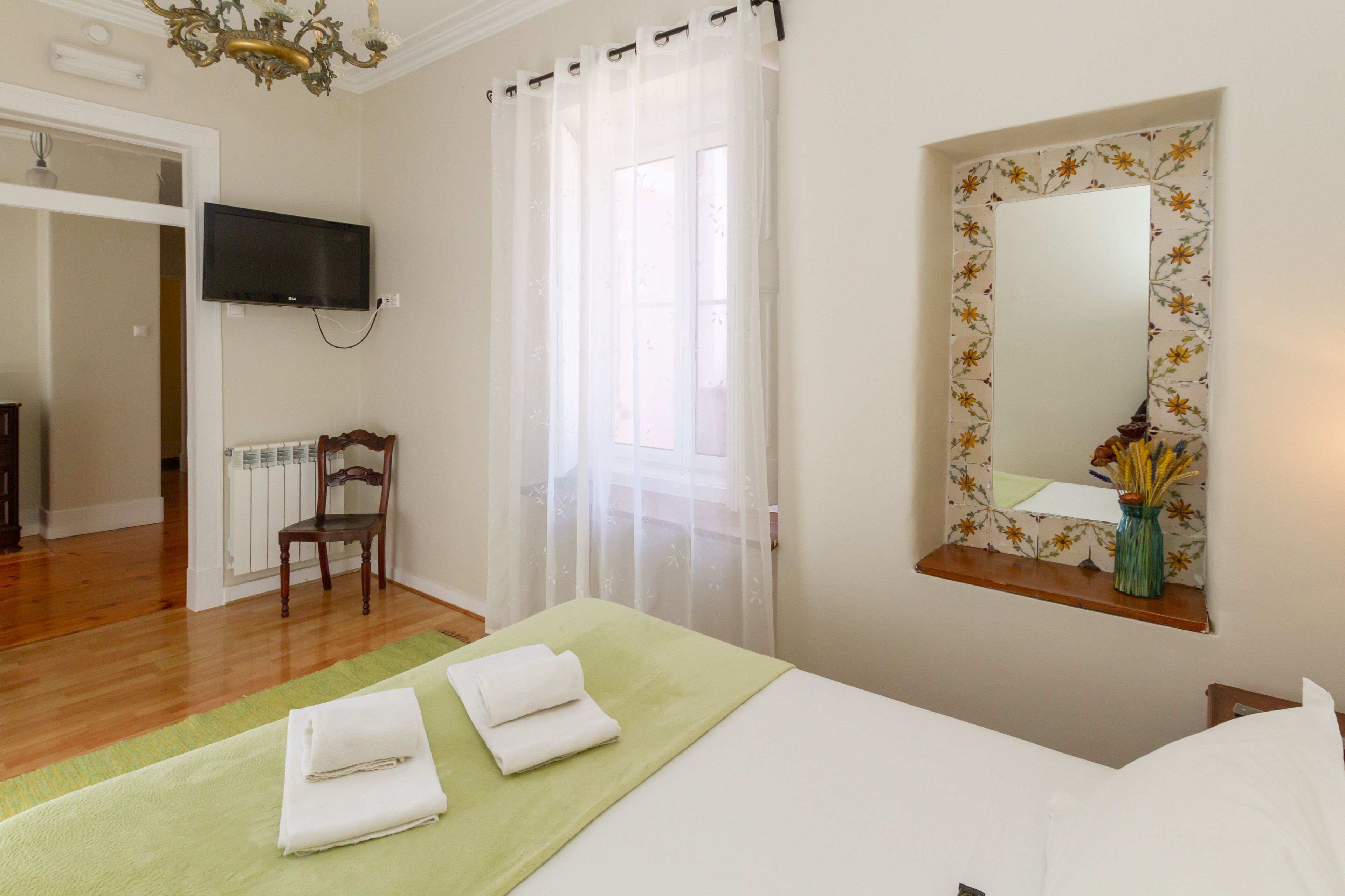 A hotel room with a bed, a television mounted on the wall, a window with sheer curtains, a mirror with a floral frame, and a radiator.
