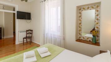 A hotel room with a bed, a television mounted on the wall, a window with sheer curtains, a mirror with a floral frame, and a radiator.
