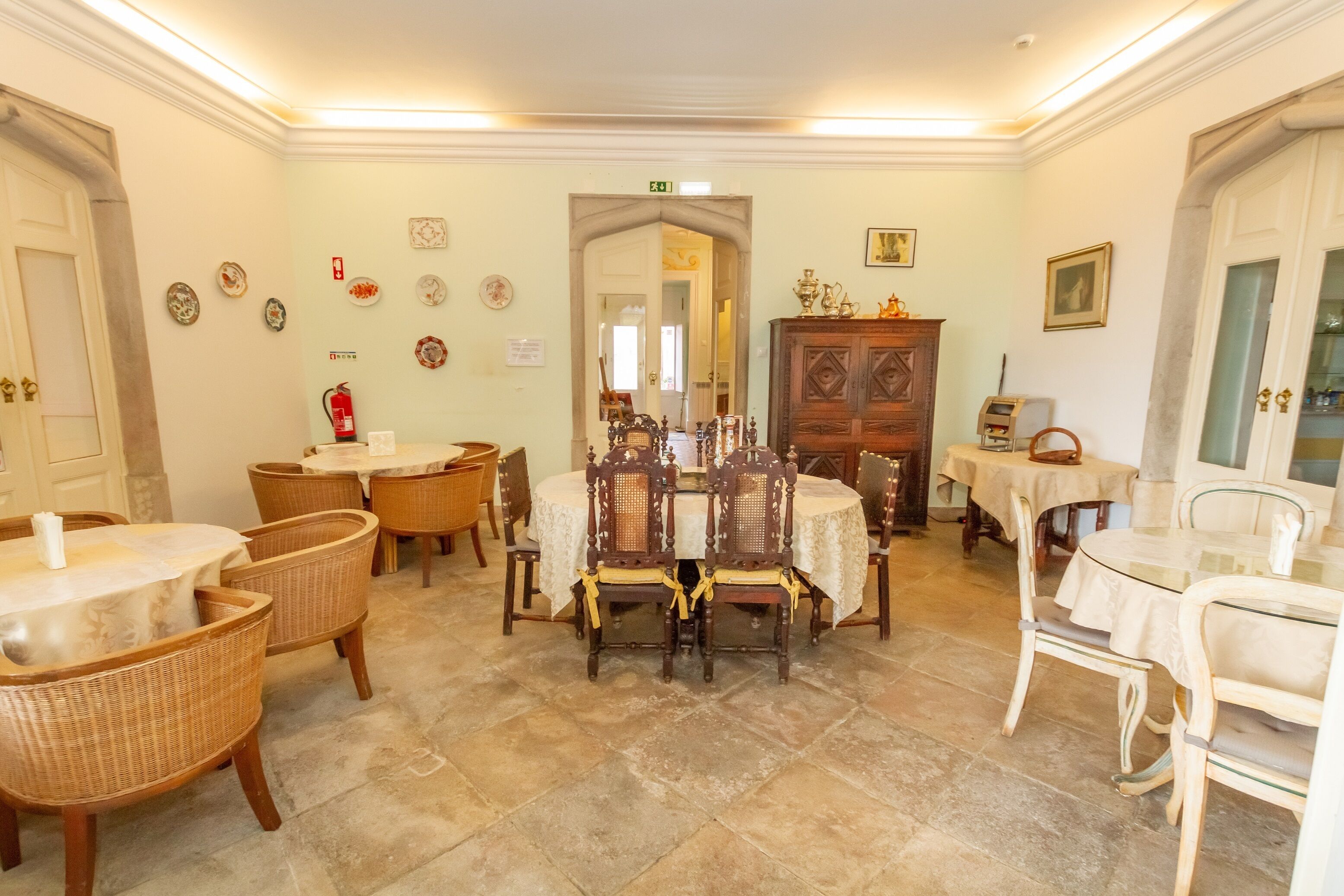 Un espace repas avec des tables rondes et des chaises en osier, un buffet en bois agrémenté d’objets décoratifs, et un extincteur fixé au mur.