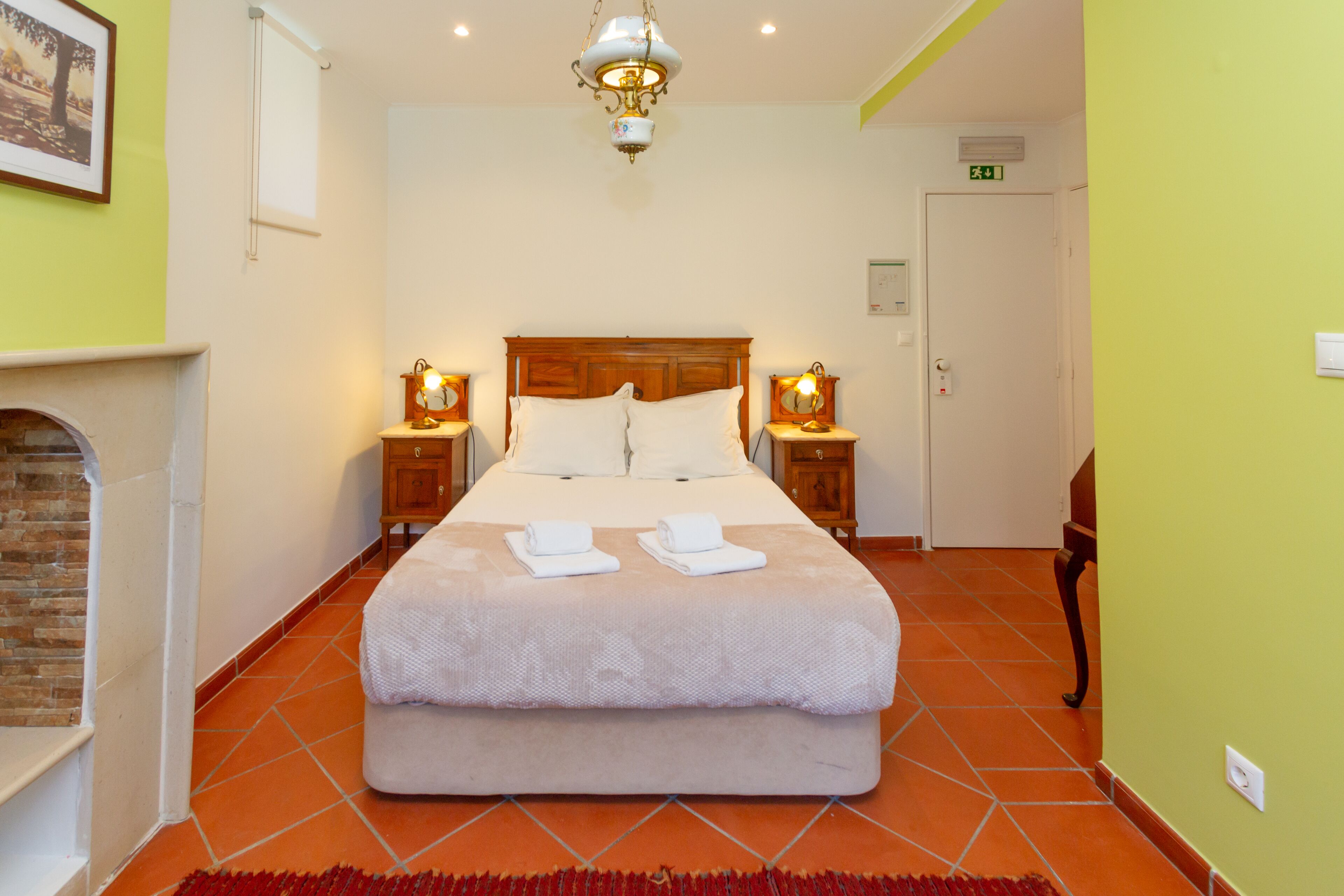 Cama bem arrumada com duas almofadas e duas toalhas dobradas, cabeceira de madeira, duas mesas de cabeceira com luminárias, lareira e piso ladrilhado.