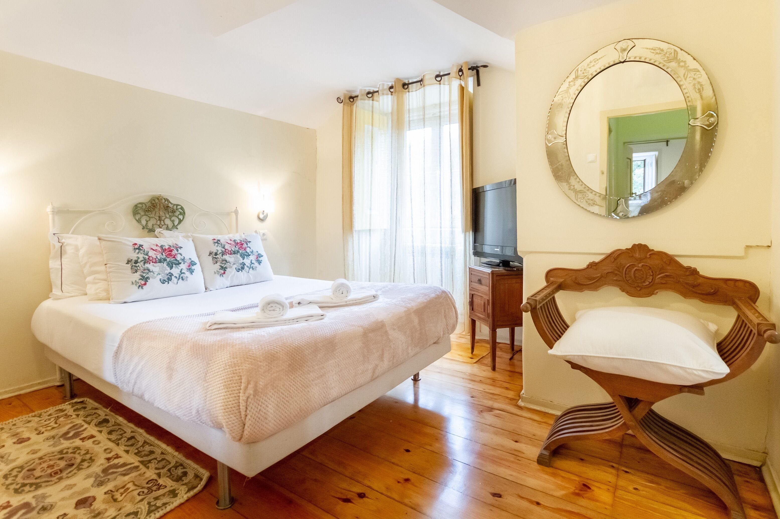 A neatly made bed with floral pillows, a wooden chair, a television on a wooden stand, and a mirror on the wall.