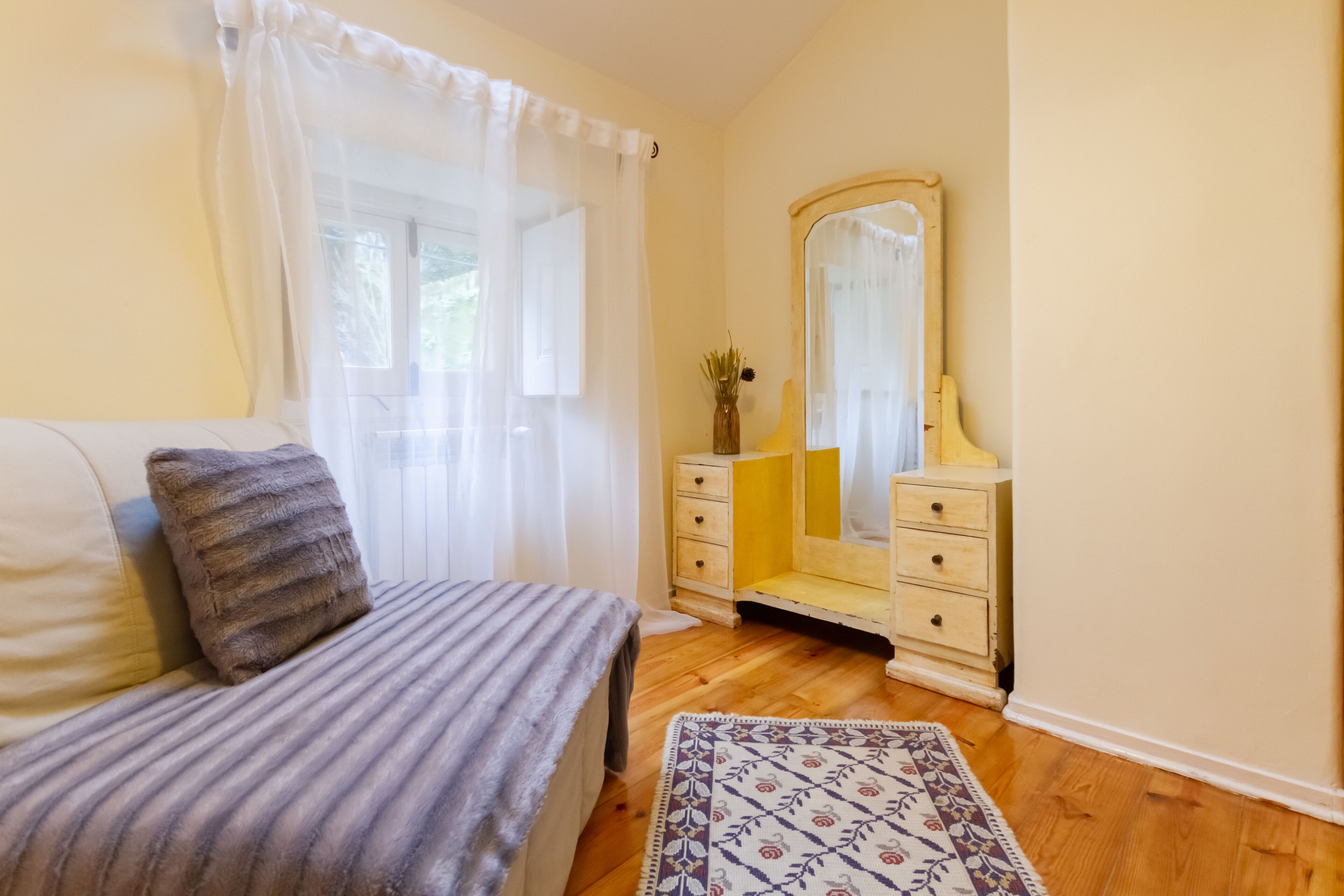Une chambre à coucher comprenant un lit, une commode, un miroir et une fenêtre ornée de rideaux légers.