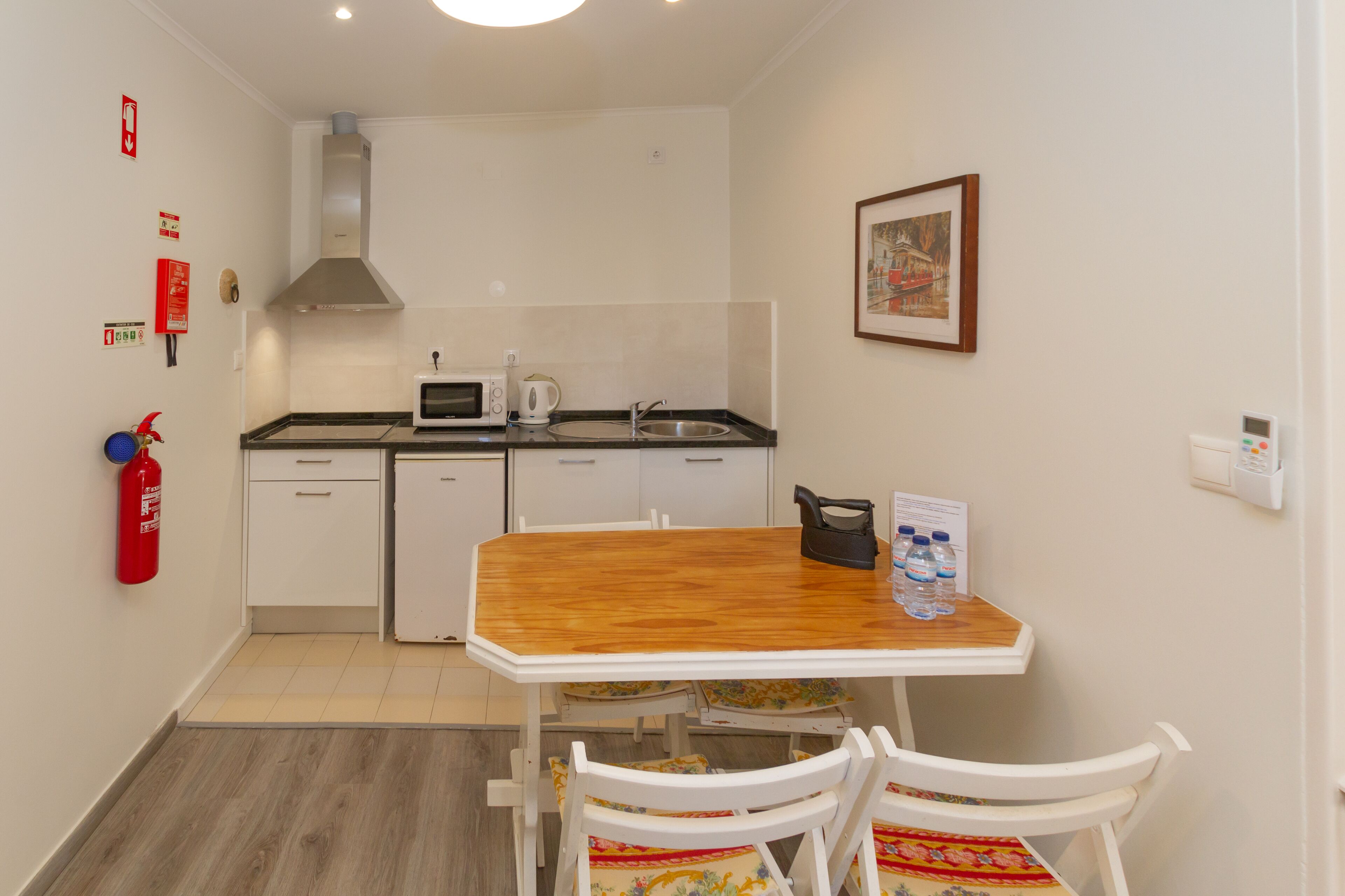 Una cocina pequeña con armarios blancos, una mesa de madera y sillas blancas. Hay un extintor en la pared y un cuadro enmarcado sobre el fregadero.
