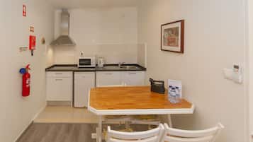 Una cocina pequeña con armarios blancos, una mesa de madera y sillas blancas. Hay un extintor en la pared y un cuadro enmarcado sobre el fregadero.