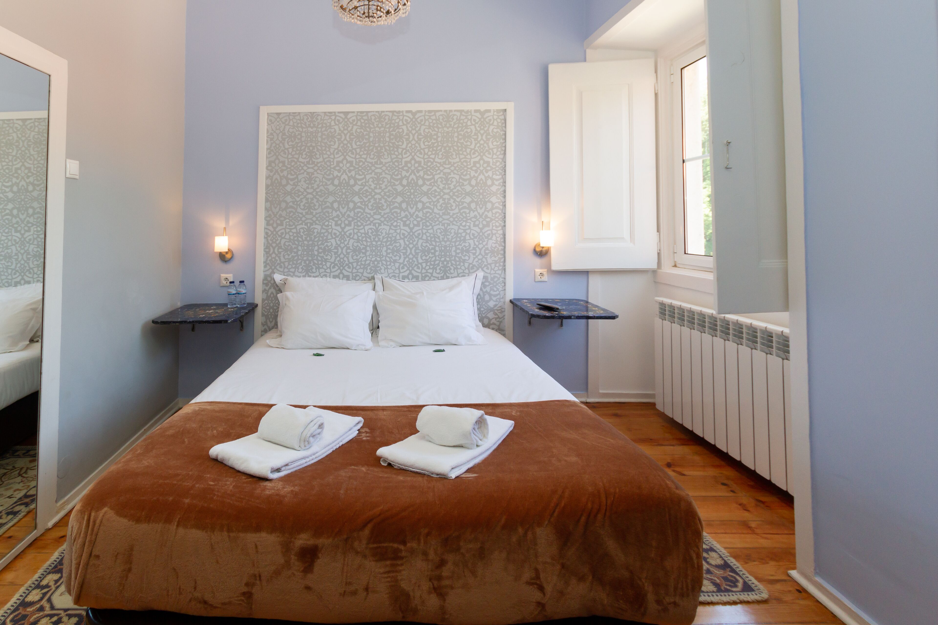 A neatly made bed with a brown cover, two bedside tables with lamps, a large window, and a radiator.