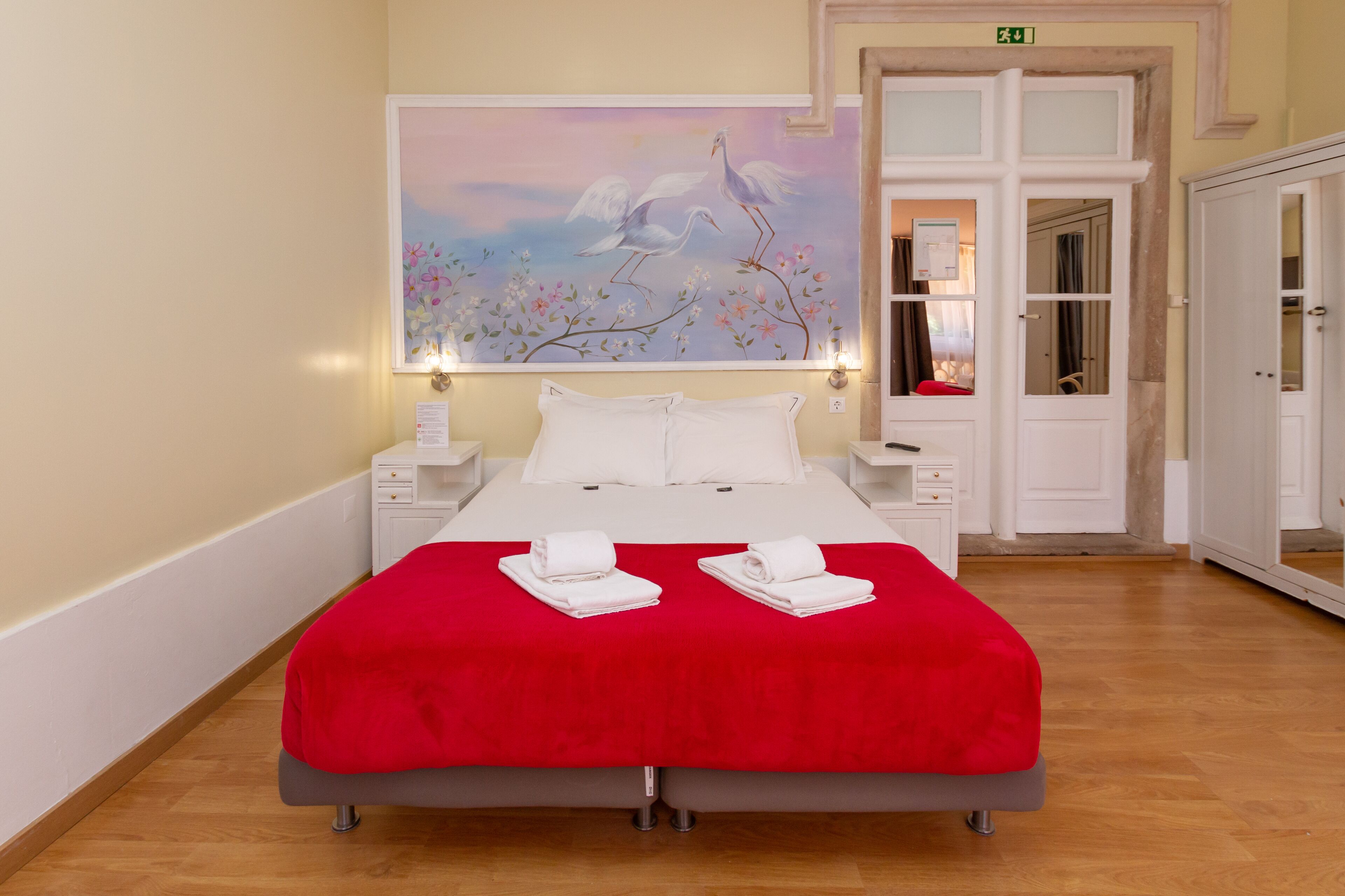 Habitación de hotel con una cama grande, una colcha roja, almohadas blancas y dos toallas dobladas con cuidado sobre la cama. En la pared, encima de la cama, hay un cuadro de pájaros.