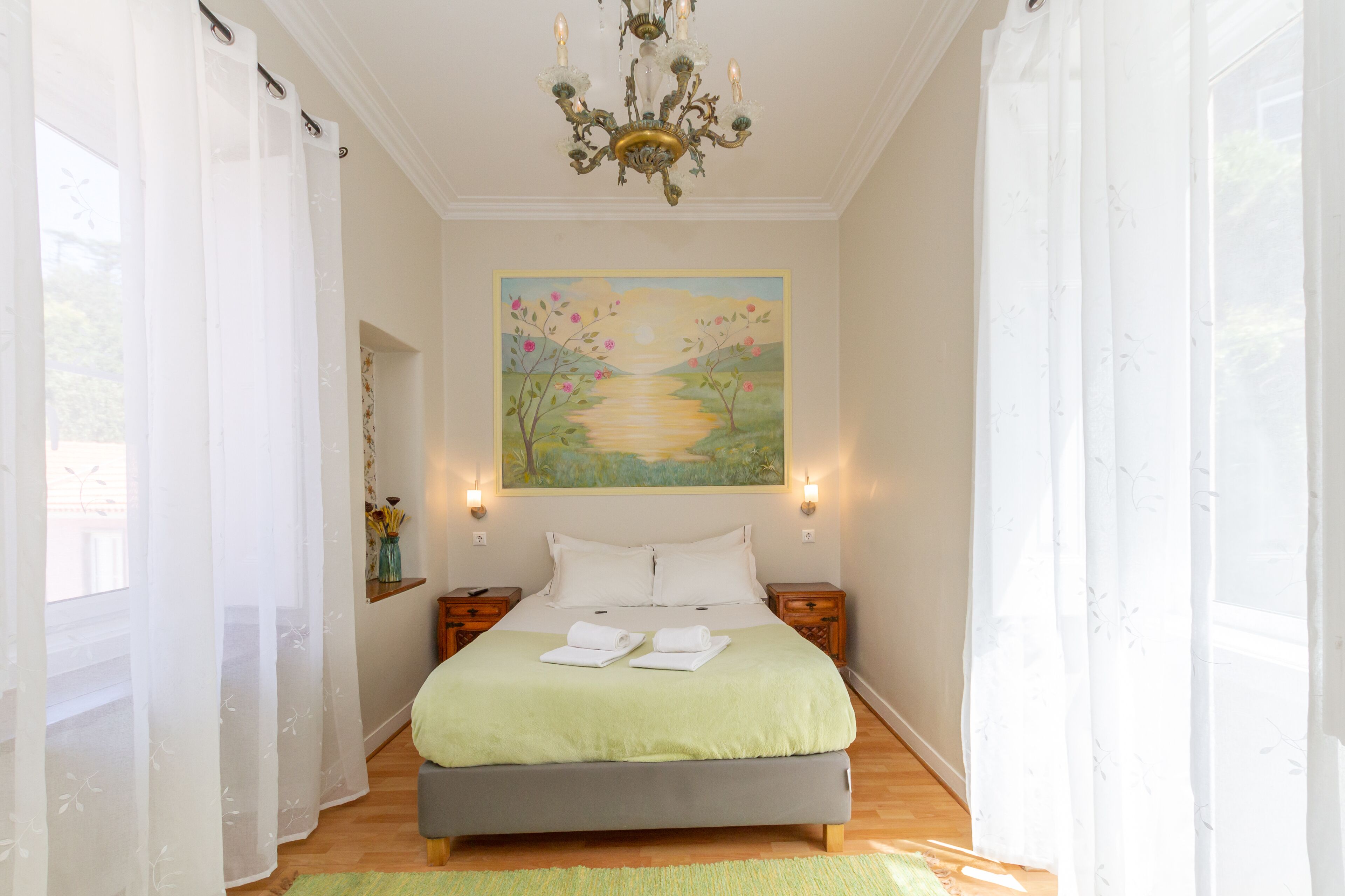 A bedroom with a large bed, two bedside tables, a chandelier, and a painting on the wall.