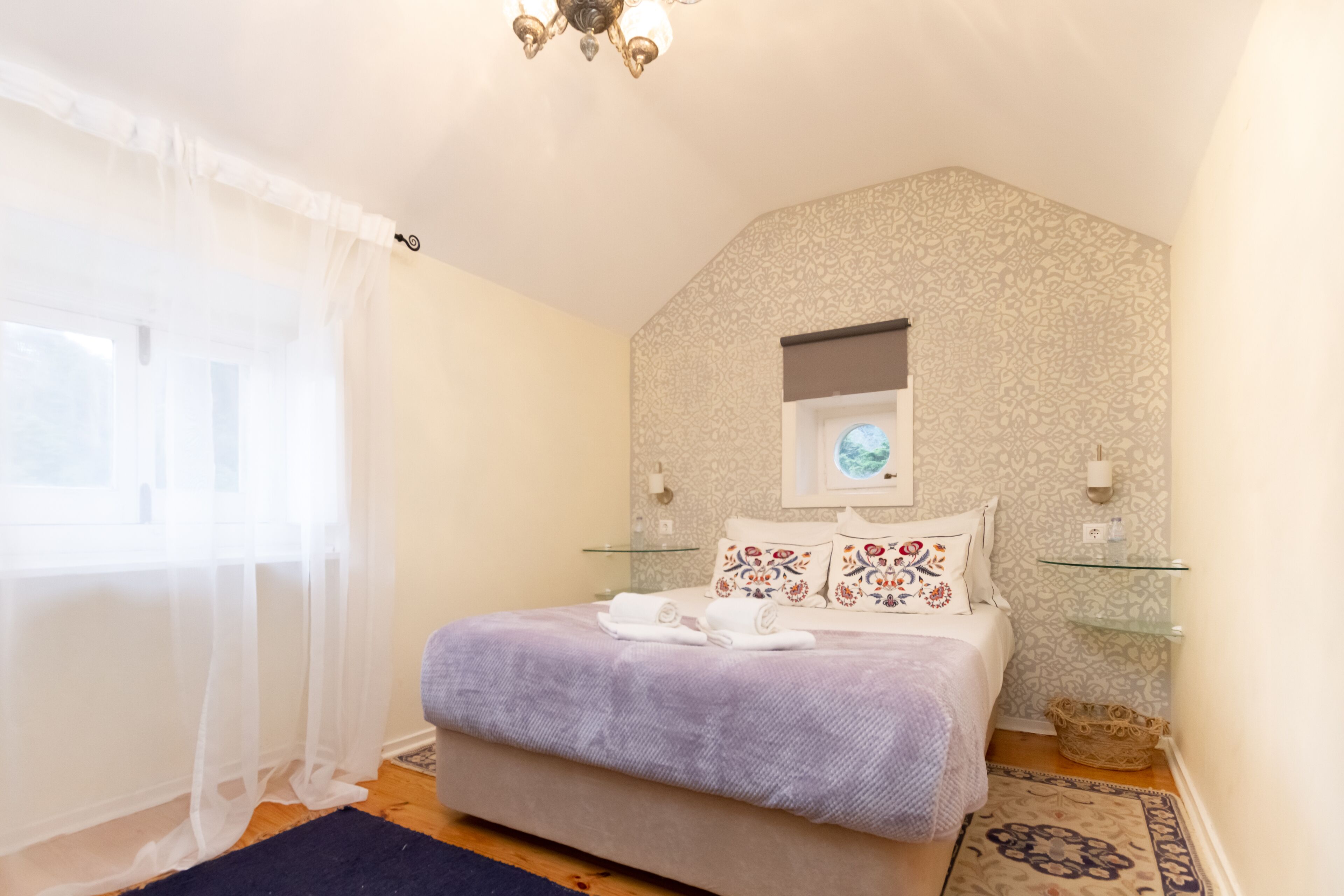 A bedroom with a large bed, a window with sheer curtains, a chandelier, and patterned wallpaper.
