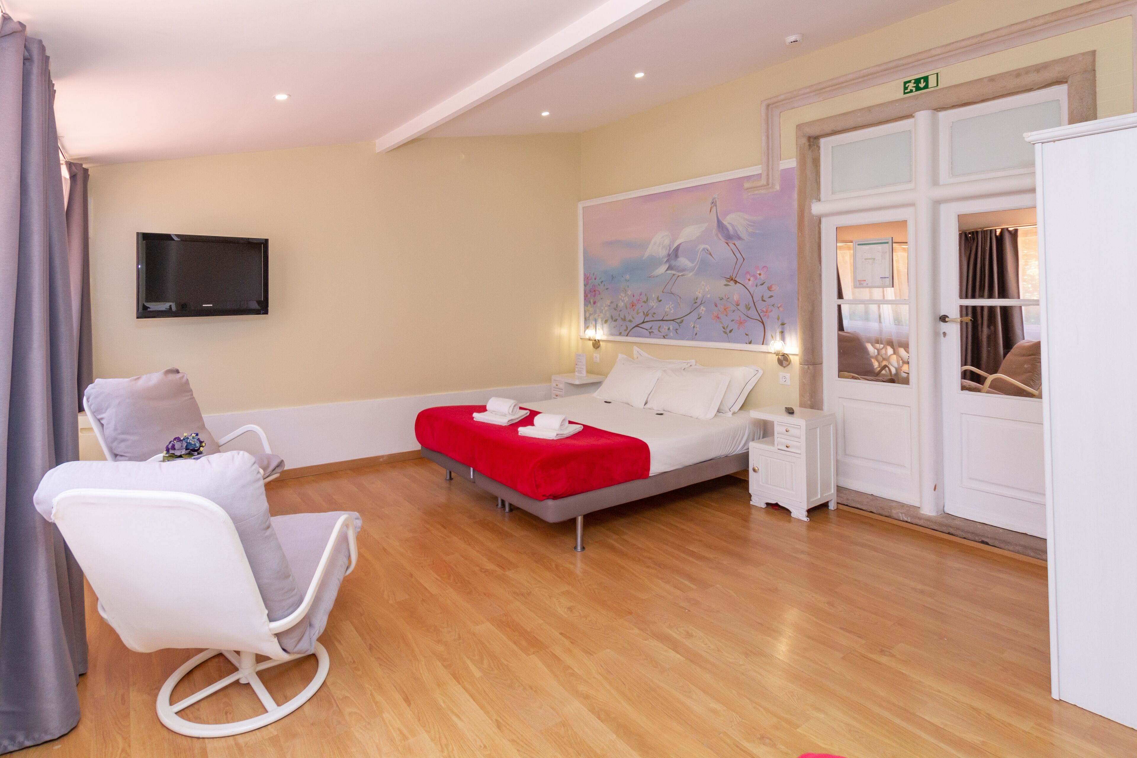 Ein Hotelzimmer mit Bett, einem weißen Sessel, einem kleinen Nachttisch, einem an der Wand montierten Fernseher und einem Bild über dem Bett.