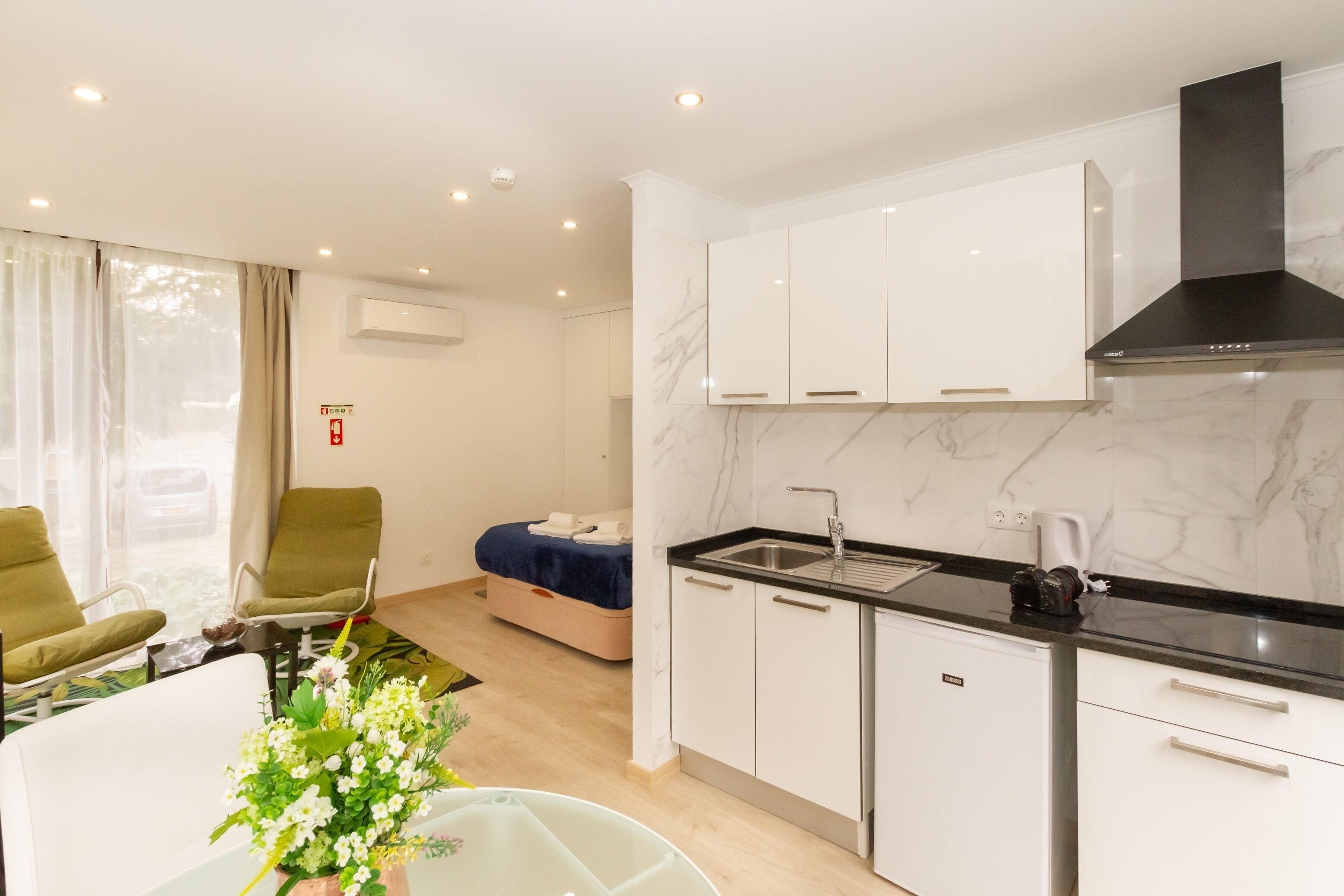 A modern kitchen with white cabinets, a black countertop, and a stainless steel sink. There is a bed with a blue blanket and a small table with a vase of flowers.