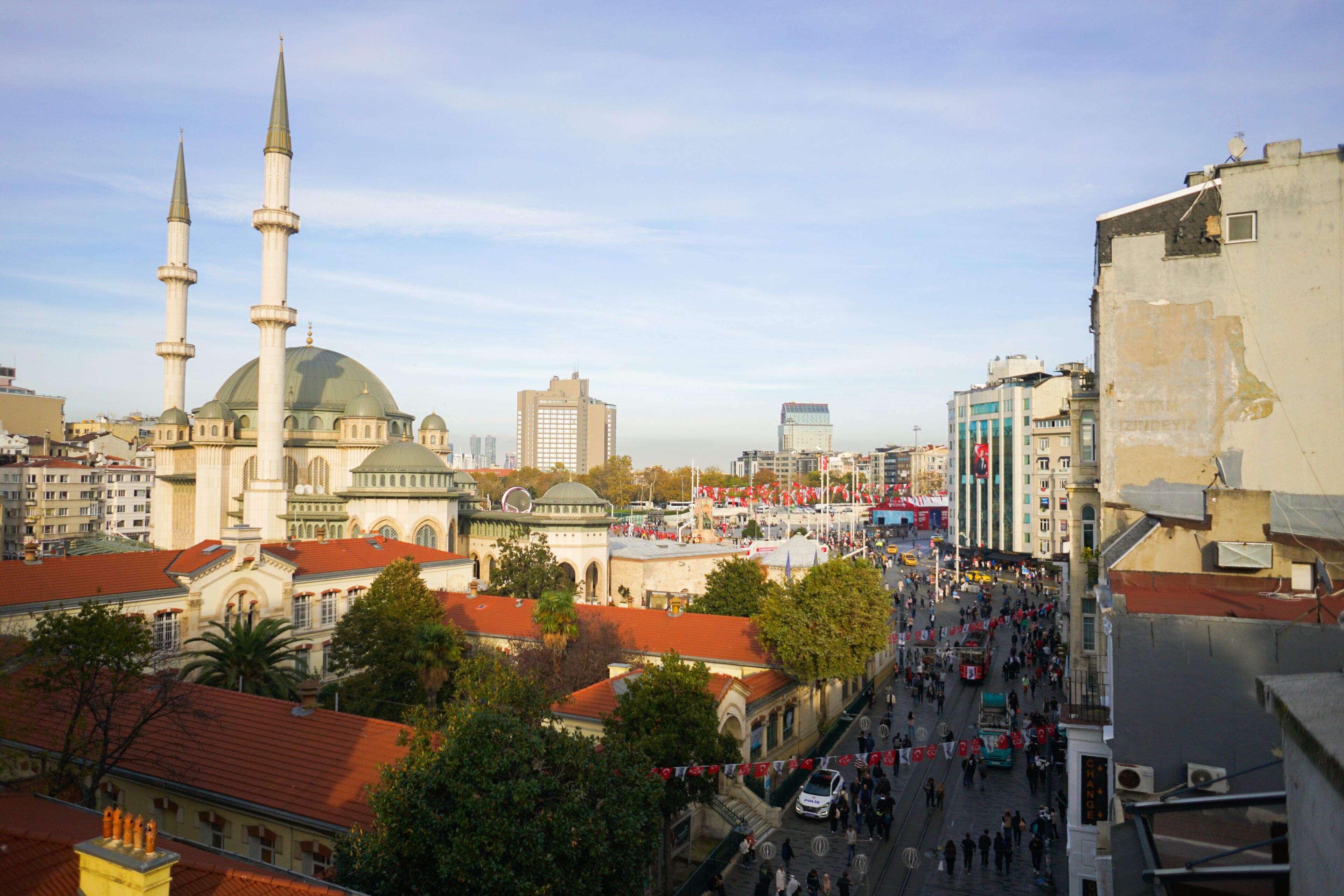 A cityscape with a prominent mosque with two minarets and a dome, surrounded by buildings and a busy street with pedestrians and vehicles.
