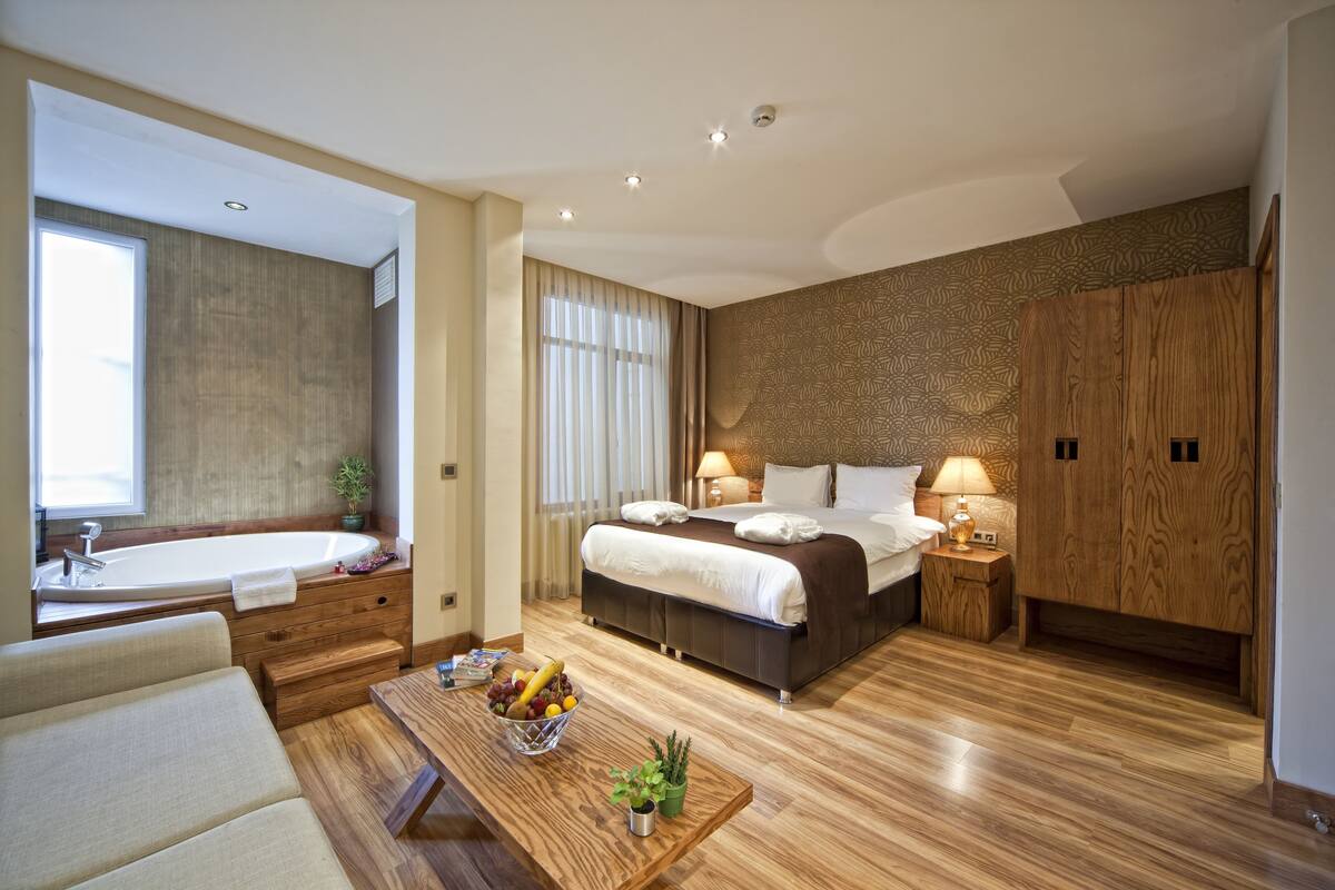 A modern hotel room with a large bed, a wooden floor, a sitting area with a table, and a bathroom with a bathtub.
