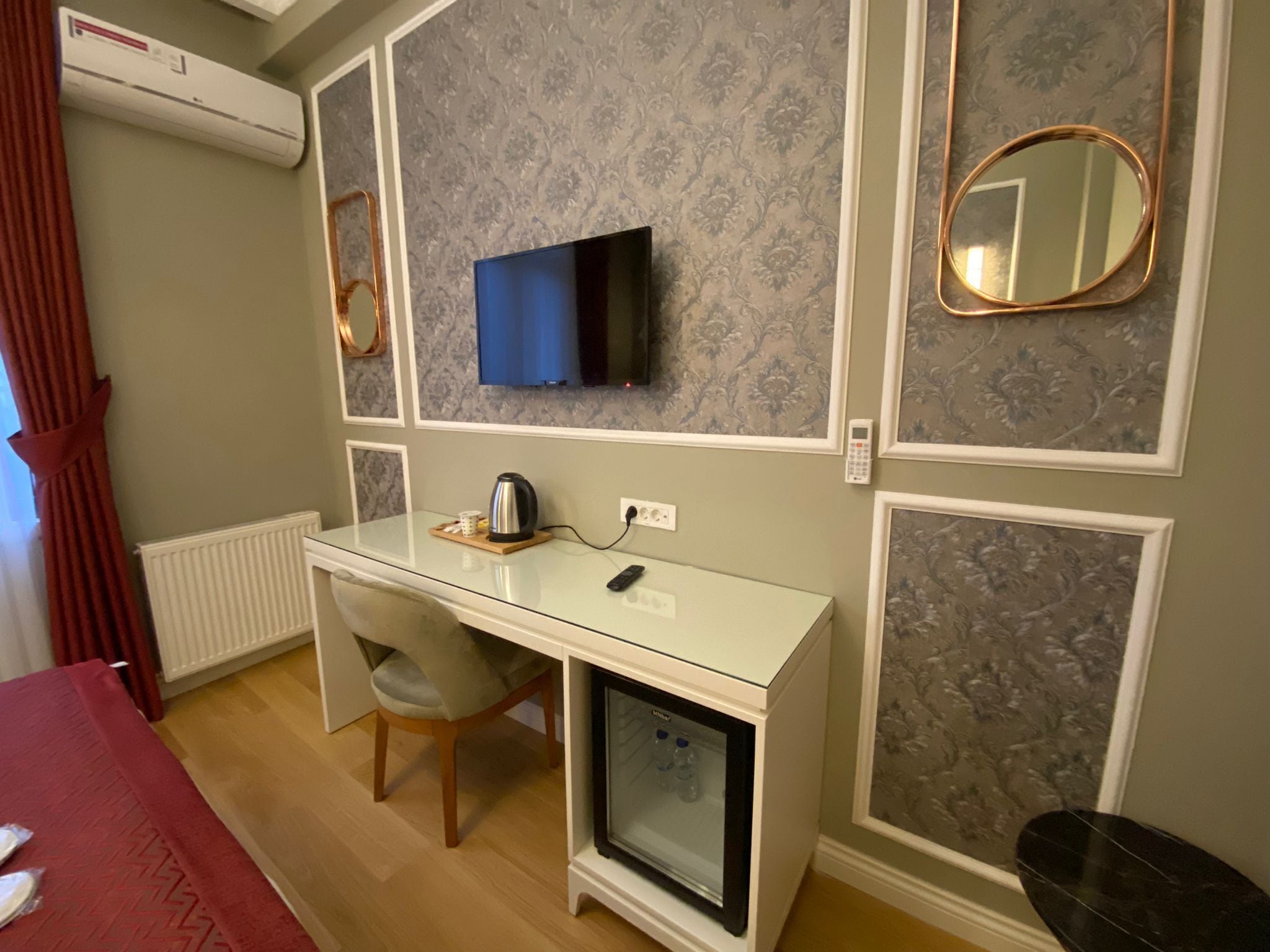 Una habitación de hotel con un escritorio blanco, un televisor montado en la pared, un refrigerador pequeño y un espejo.