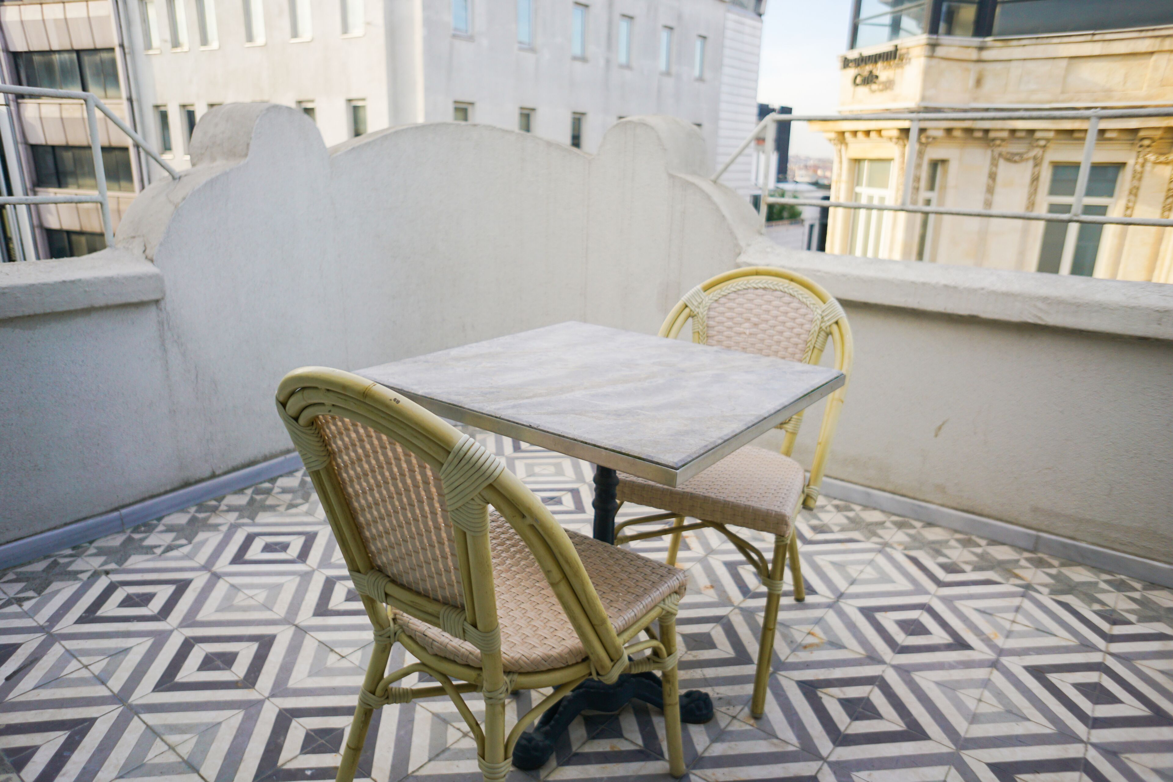 A balcony with two wicker chairs and a table, patterned floor tiles, and a white wall with a railing.