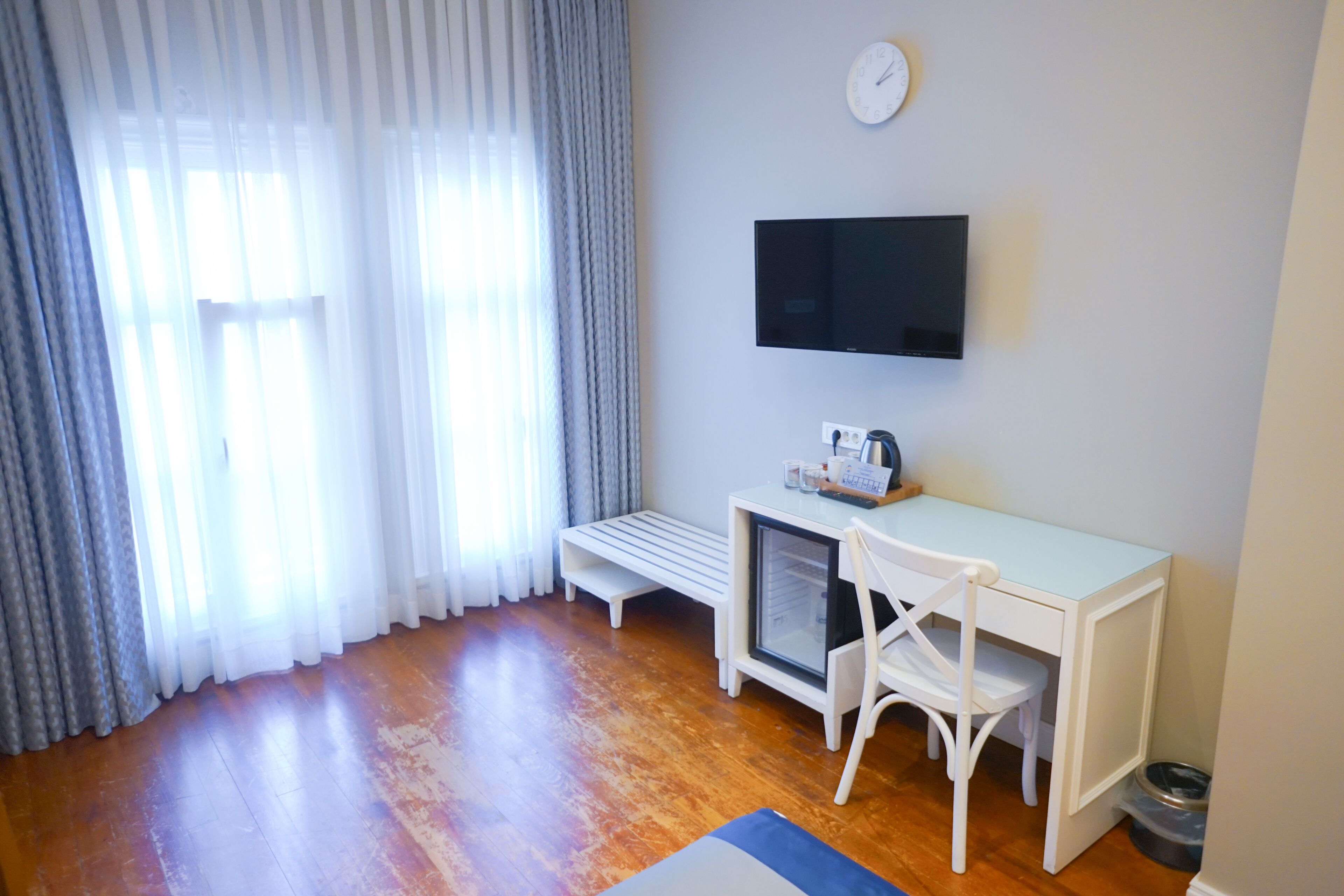 A room with a wooden floor, a white desk with a chair, a television mounted on the wall, and a window with sheer curtains.
