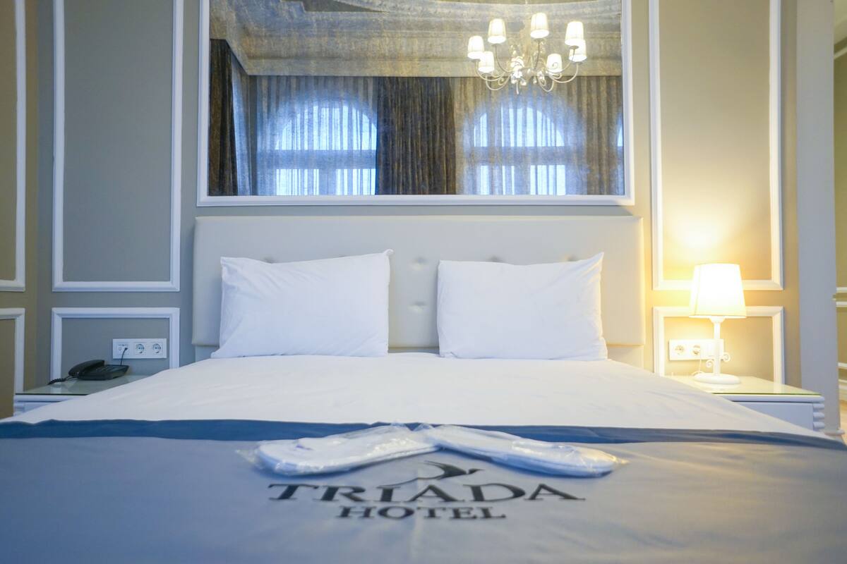 A neatly made bed with white linens, a chandelier above, and a window with curtains.