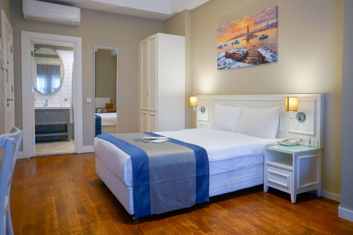 A hotel room with a bed, bedside tables, a mirror, a painting, and a bathroom visible through an open door.