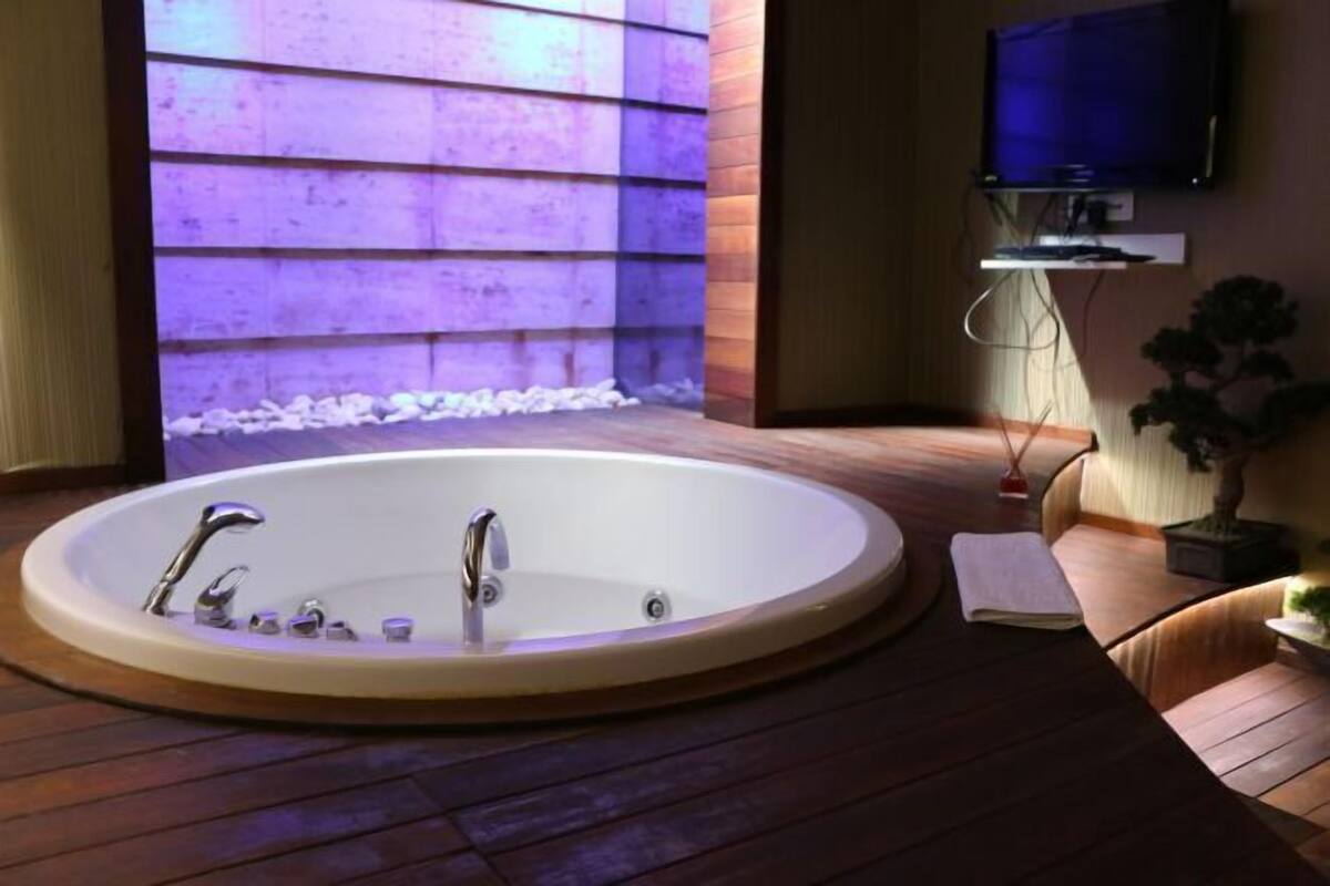 A modern bathroom with a freestanding bathtub, wooden flooring, a television mounted on the wall, and a potted plant.
