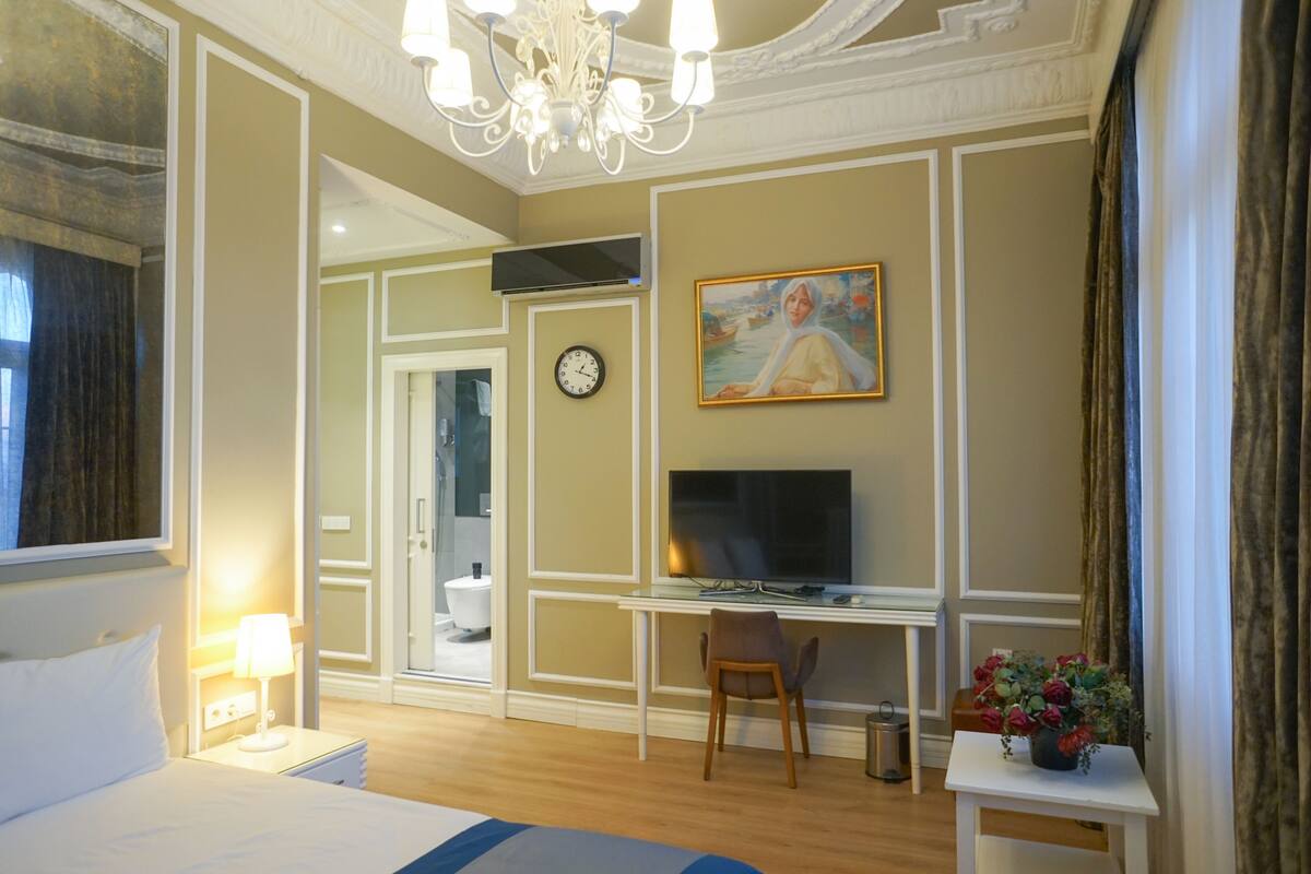 A hotel room with a bed, desk, chair, TV, clock, and a painting on the wall.