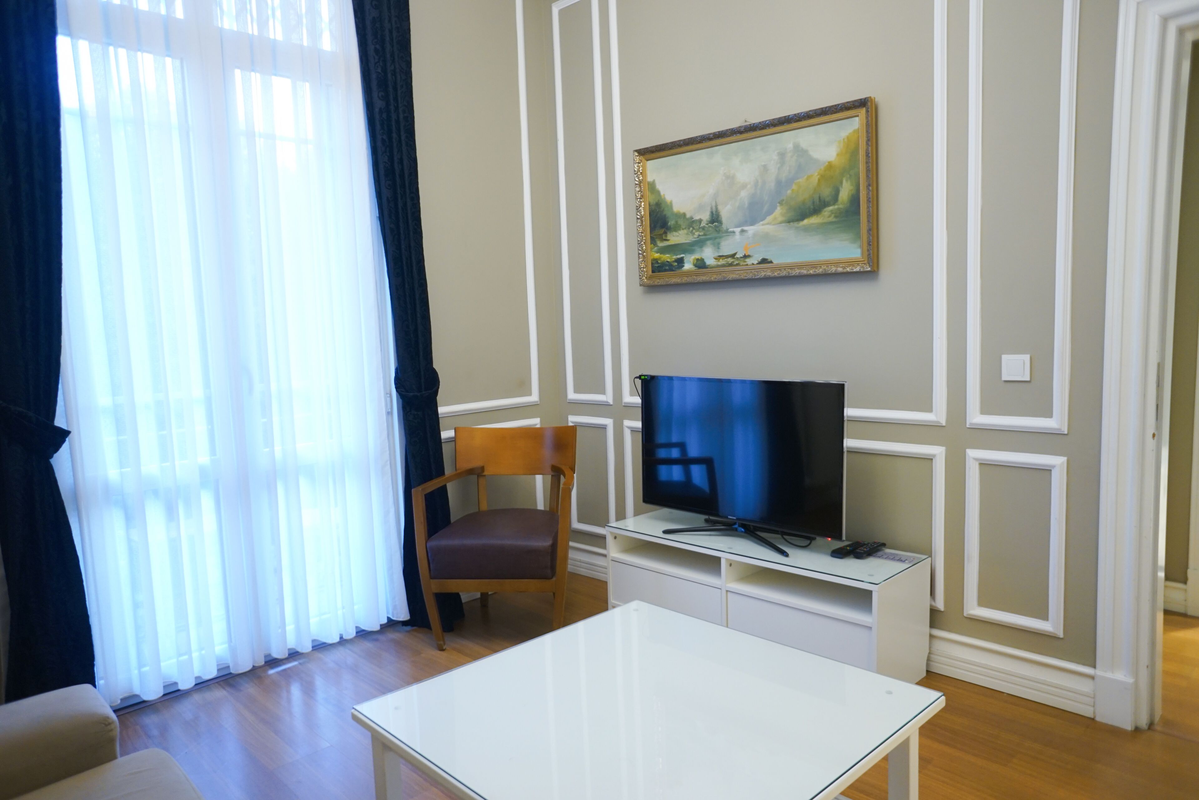 A living room with a flat-screen TV on a stand, a wooden chair, a white coffee table, and a painting on the wall.