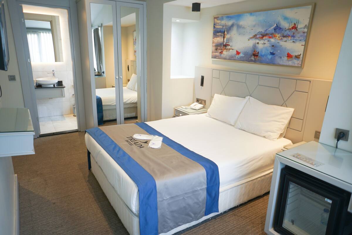 A hotel room with a bed, a bedside table, a nightstand, a mirror, a painting on the wall, and a bathroom visible through an open door.