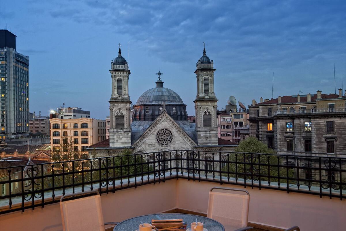 A terrace with a round table and four chairs, overlooking a historic building with two towers and a dome.