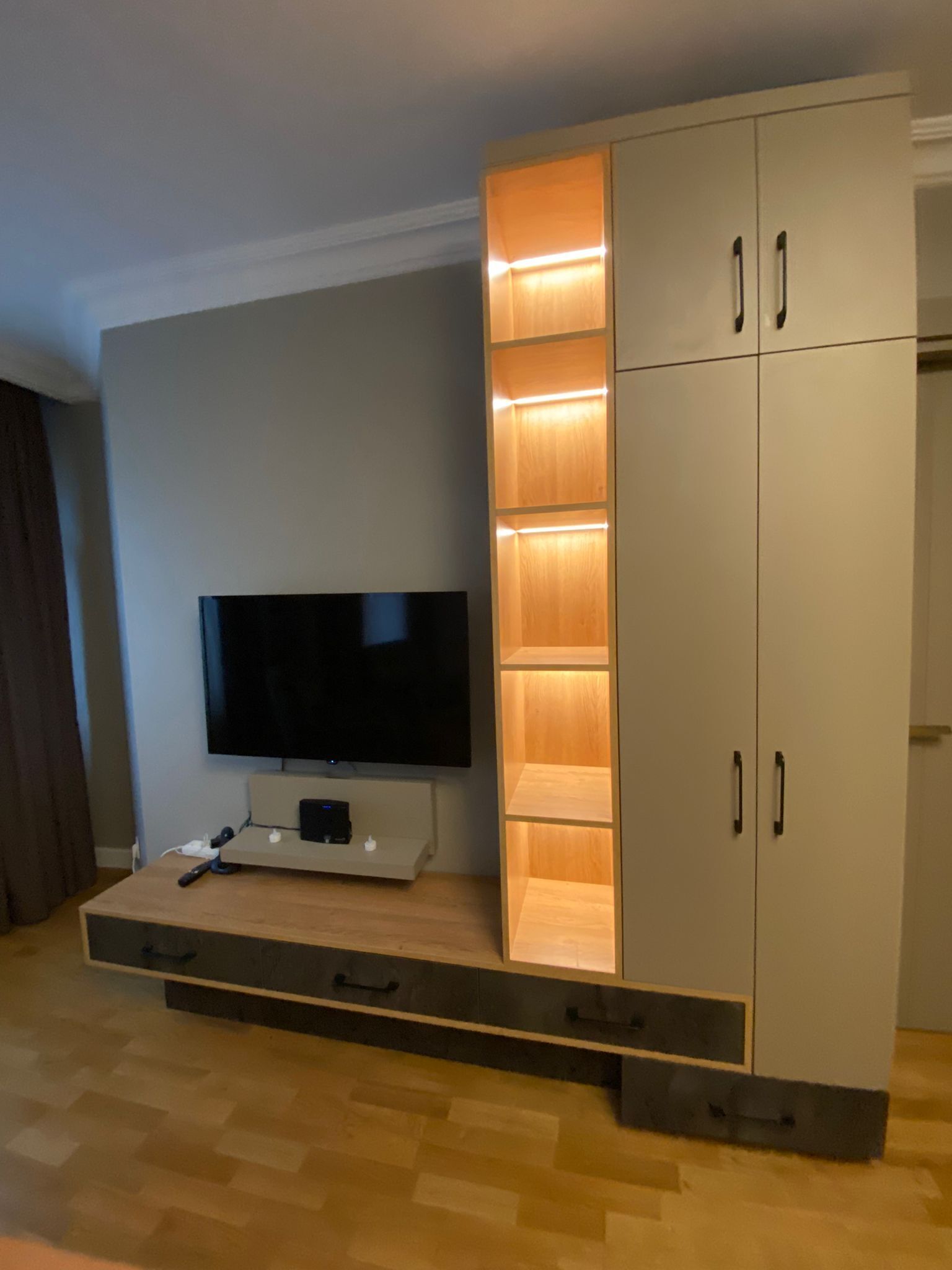 A modern living room with a flat-screen TV mounted on a wooden platform, a large cabinet with illuminated shelves, and a wooden floor.