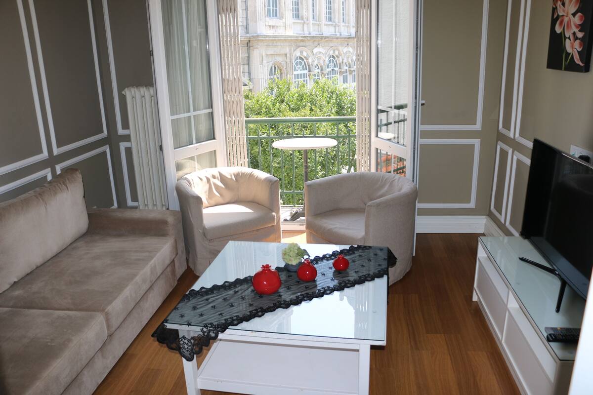 A living room with a sofa, two armchairs, a glass coffee table with decorative items, a television, and a view of a balcony with greenery.
