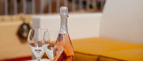 Una mesa con una botella de champagne Brut, dos copas vacías y una servilleta sobre un mantel blanco. Al fondo, un sofá amarillo y una barandilla metálica.