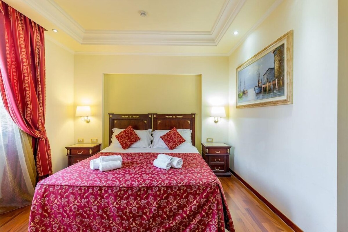 A hotel room with a large bed, two bedside tables, a painting on the wall, and red curtains.