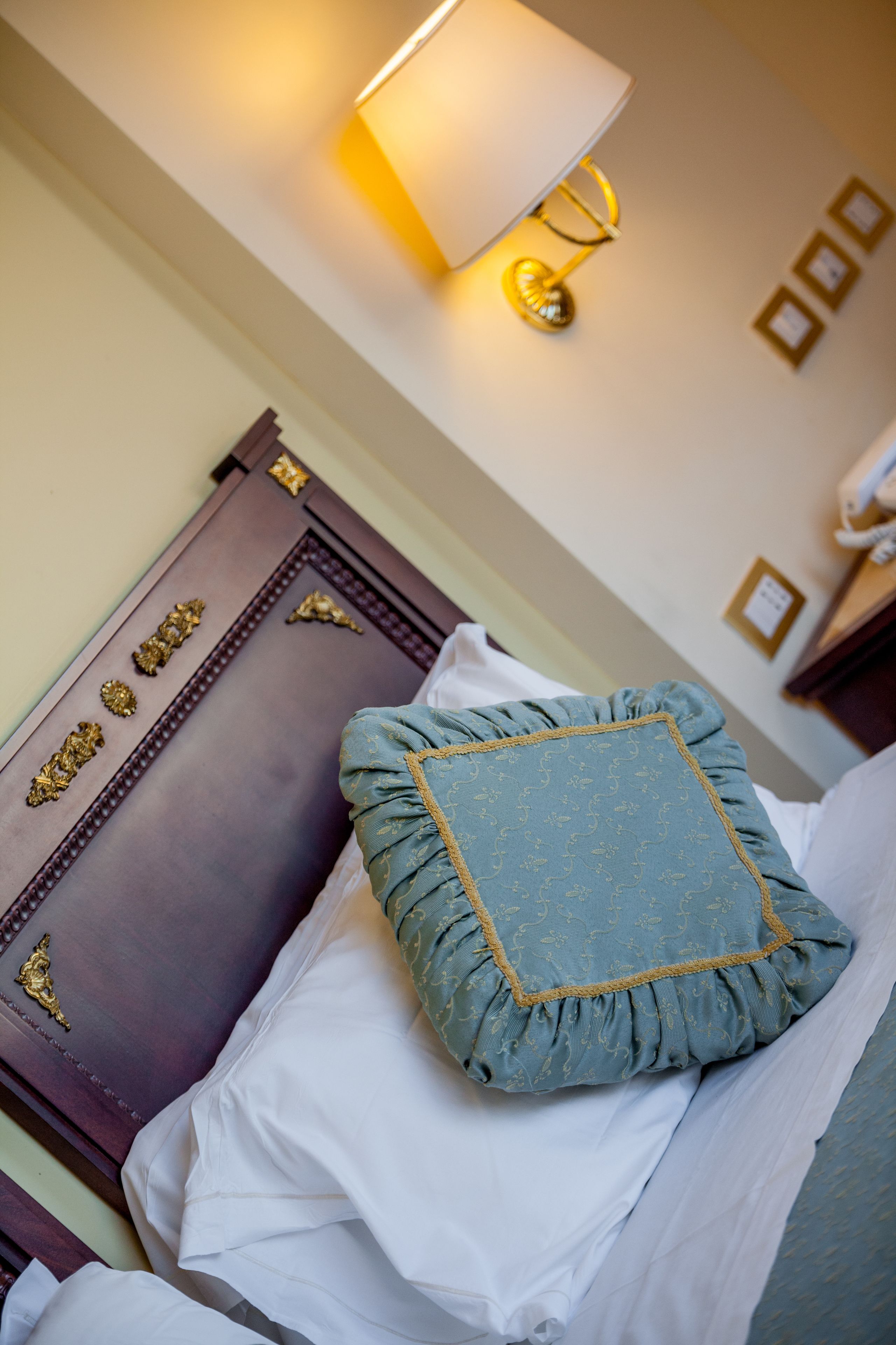 A neatly made bed with a blue pillow and white linens, a wooden headboard with decorative gold accents, and a wall-mounted lamp.
