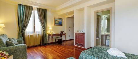 A hotel room with a wooden floor, a sofa, a bed with floral bedding, a desk with a chair, and a bathroom visible through an open door.
