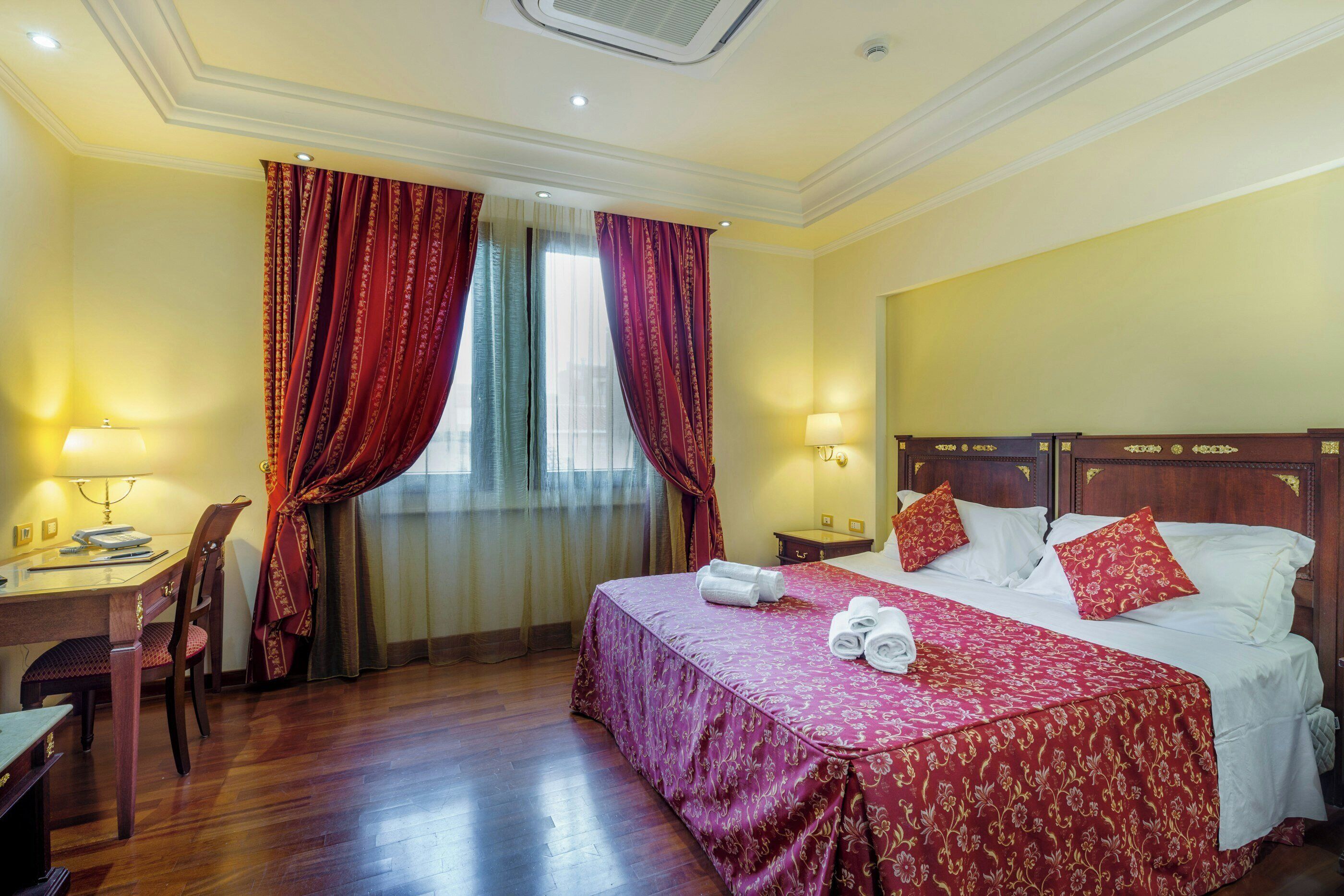 A hotel room with a wooden bed, red curtains, a desk with a lamp, and a chair.