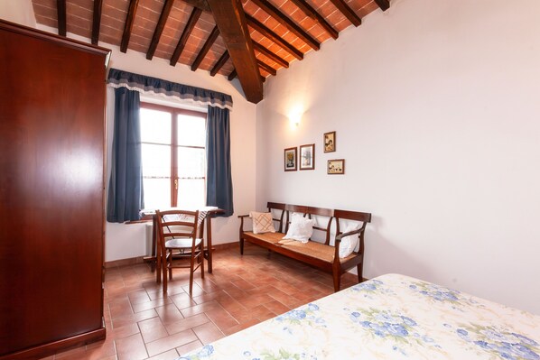 A room with a wooden wardrobe, a wooden bench, a chair, a bed with a floral bedspread, a window with blue curtains, and a wall-mounted light.