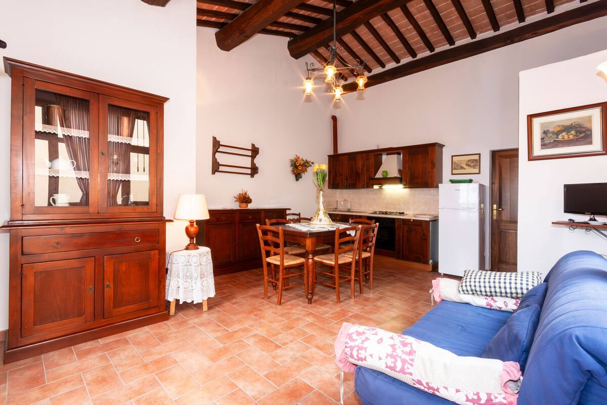 Una sala de estar acogedora con un sofá azul, una mesa de comedor de madera y una cocina con armarios de madera y suelo de baldosas.
