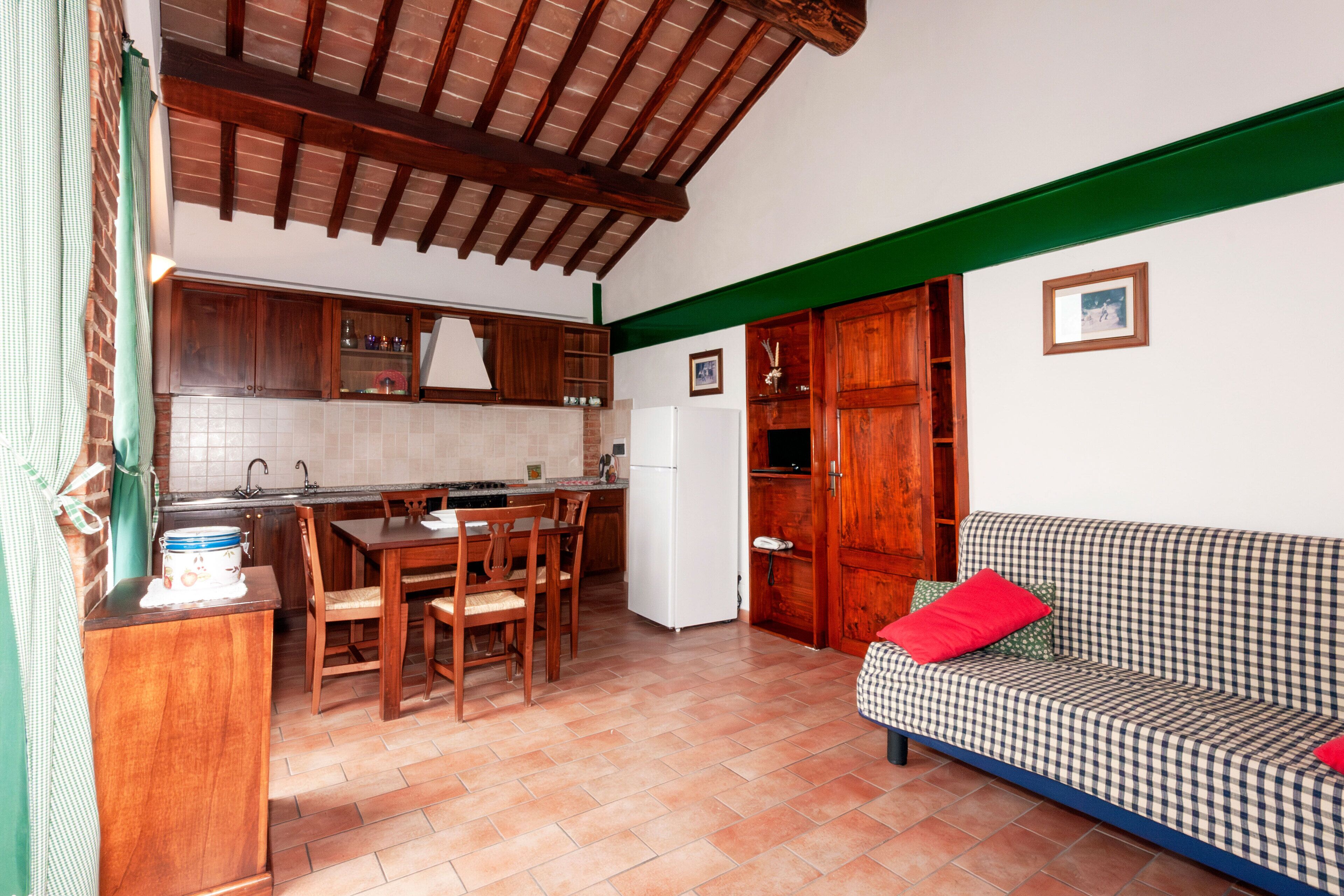 A kitchen with wooden cabinets, a dining table with chairs, a refrigerator, and a checkered sofa with red cushions.