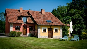 A two-story house with a red-tiled roof, a stone chimney, and a small window. There are two lounge chairs in the front yard. - La Lluriga (Llanes)