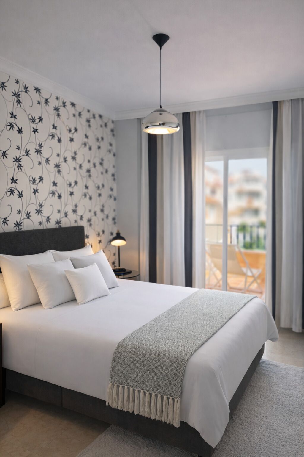 A modern bedroom with a large bed, a hanging light fixture, and a view of the outdoors.