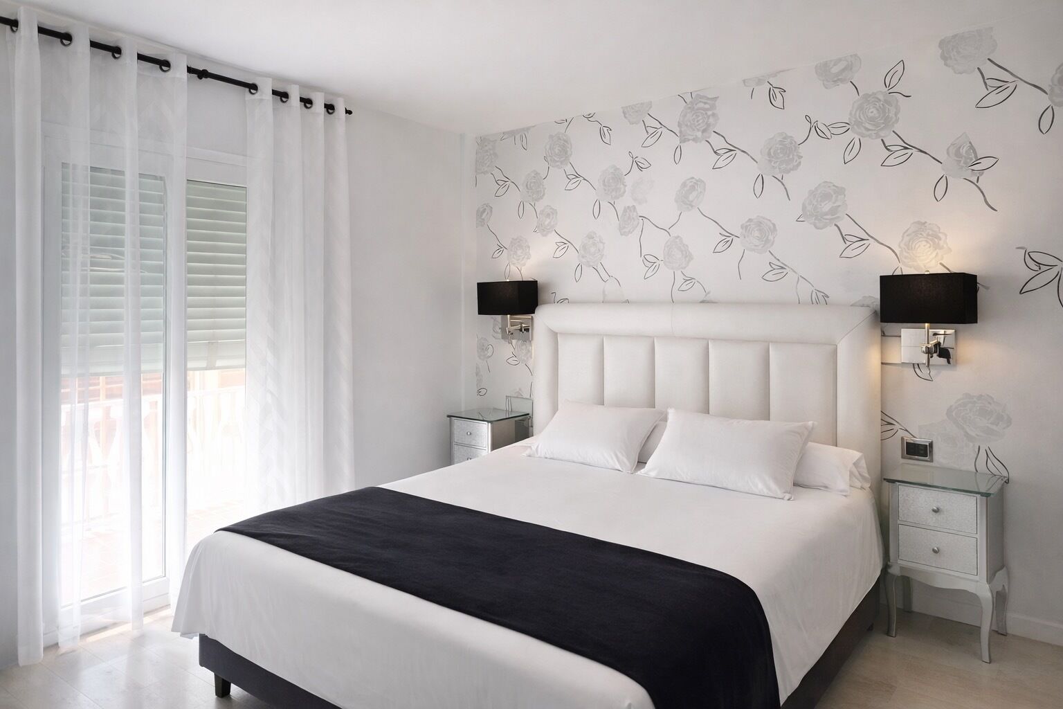 Un dormitorio con cama, cortinas blancas, una pared con motivos florales, dos lámparas de noche y una mesita de noche.