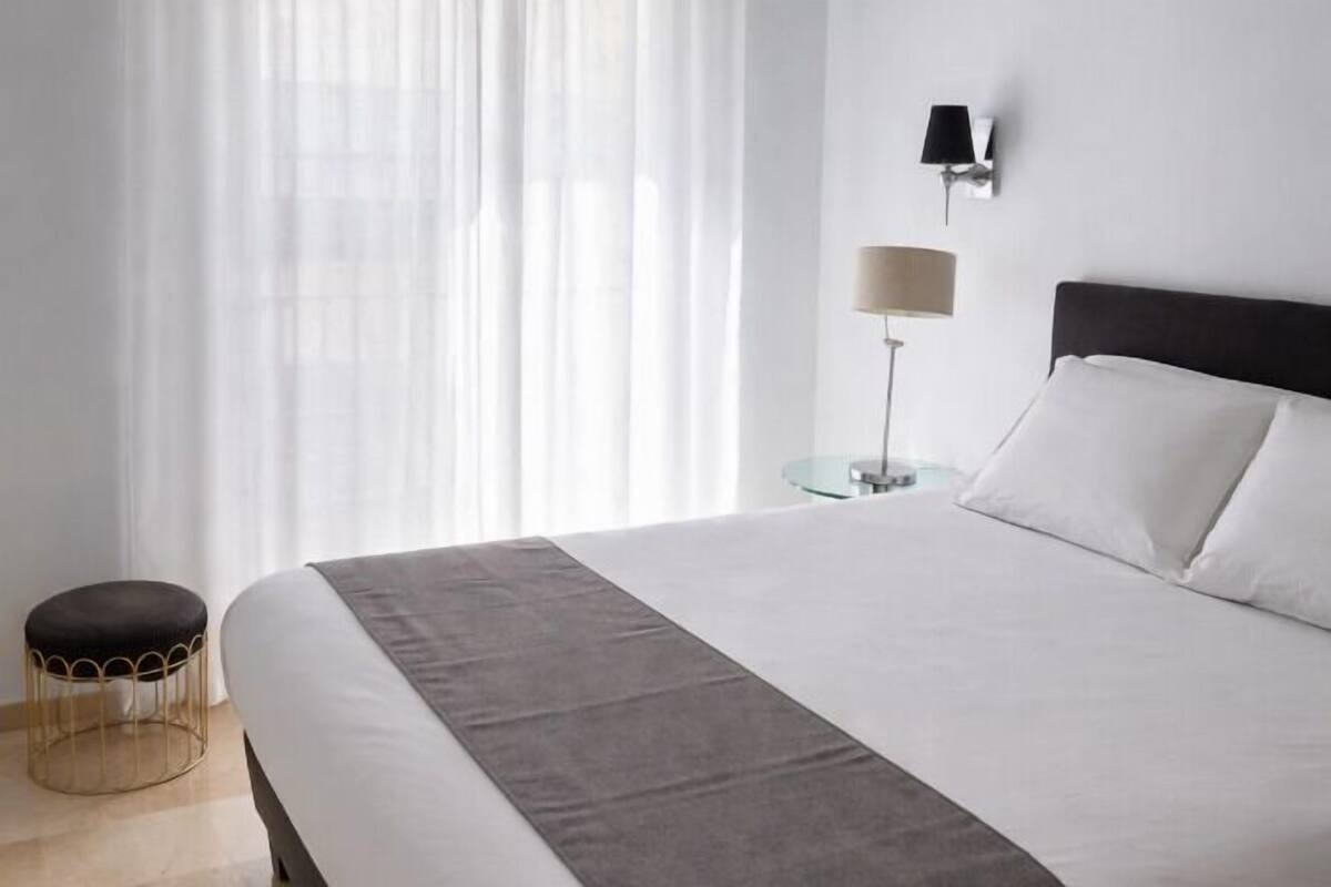 Una habitación de hotel con una cama grande, una mesita redonda pequeña, una lámpara y una ventana con cortinas traslúcidas.
