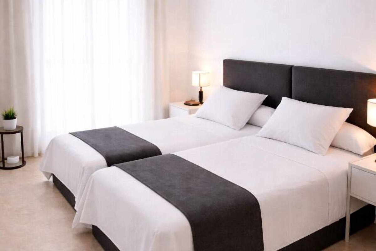 Cama doble con cabecera oscura, ropa de cama blanca y mesita de noche con lámpara.
