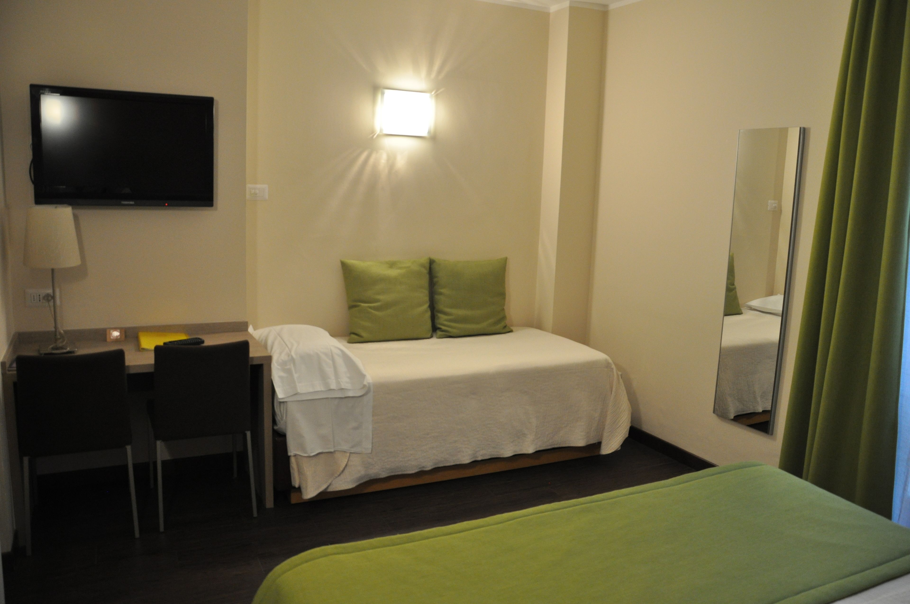 Ett hotellrum med en säng, ett skrivbord med en stol, en tv som är monterad på väggen och en spegel.